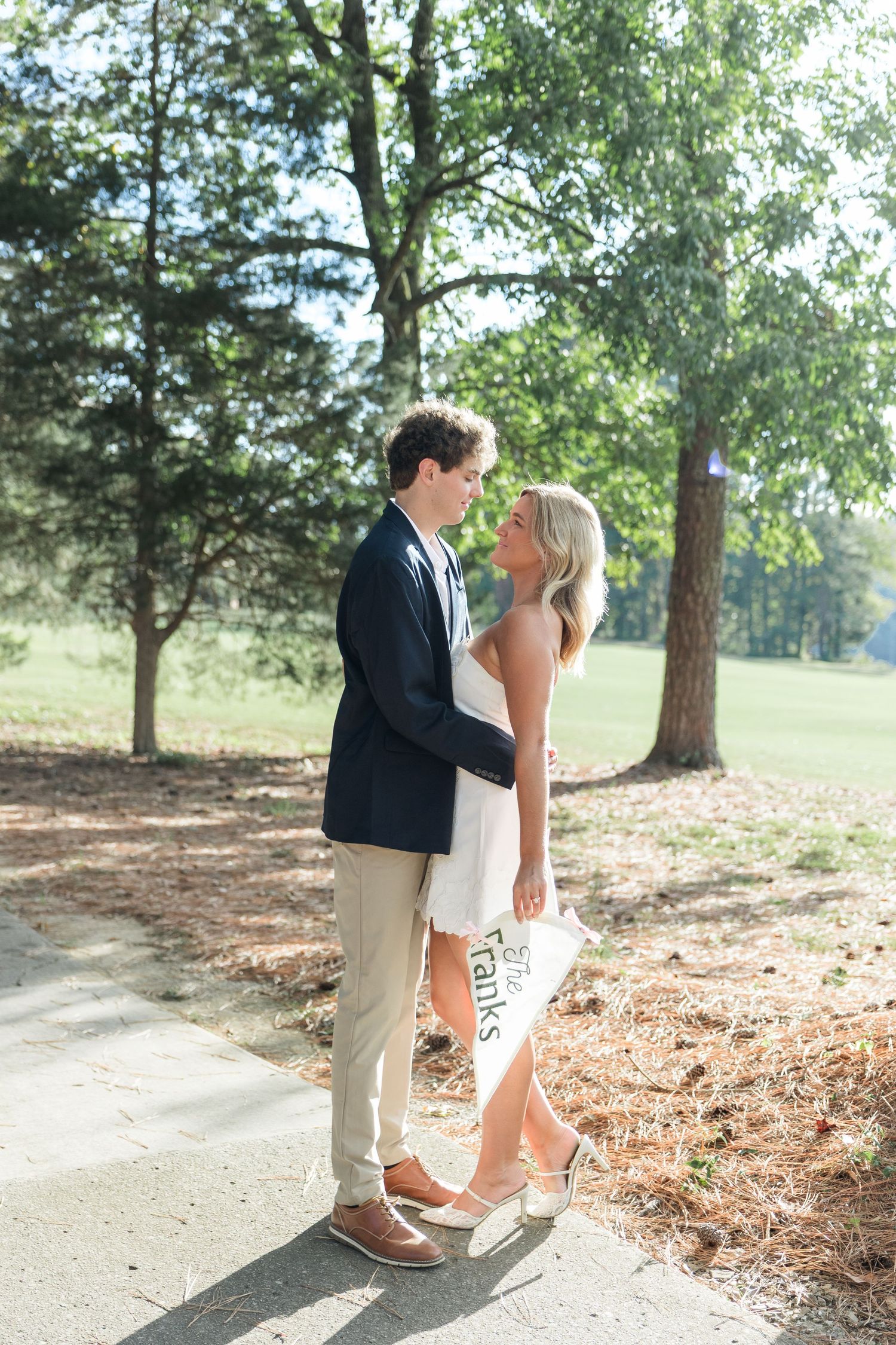 Couple sharing a romantic moment on a sunlit path, woman holding a decorative sign in white dress.