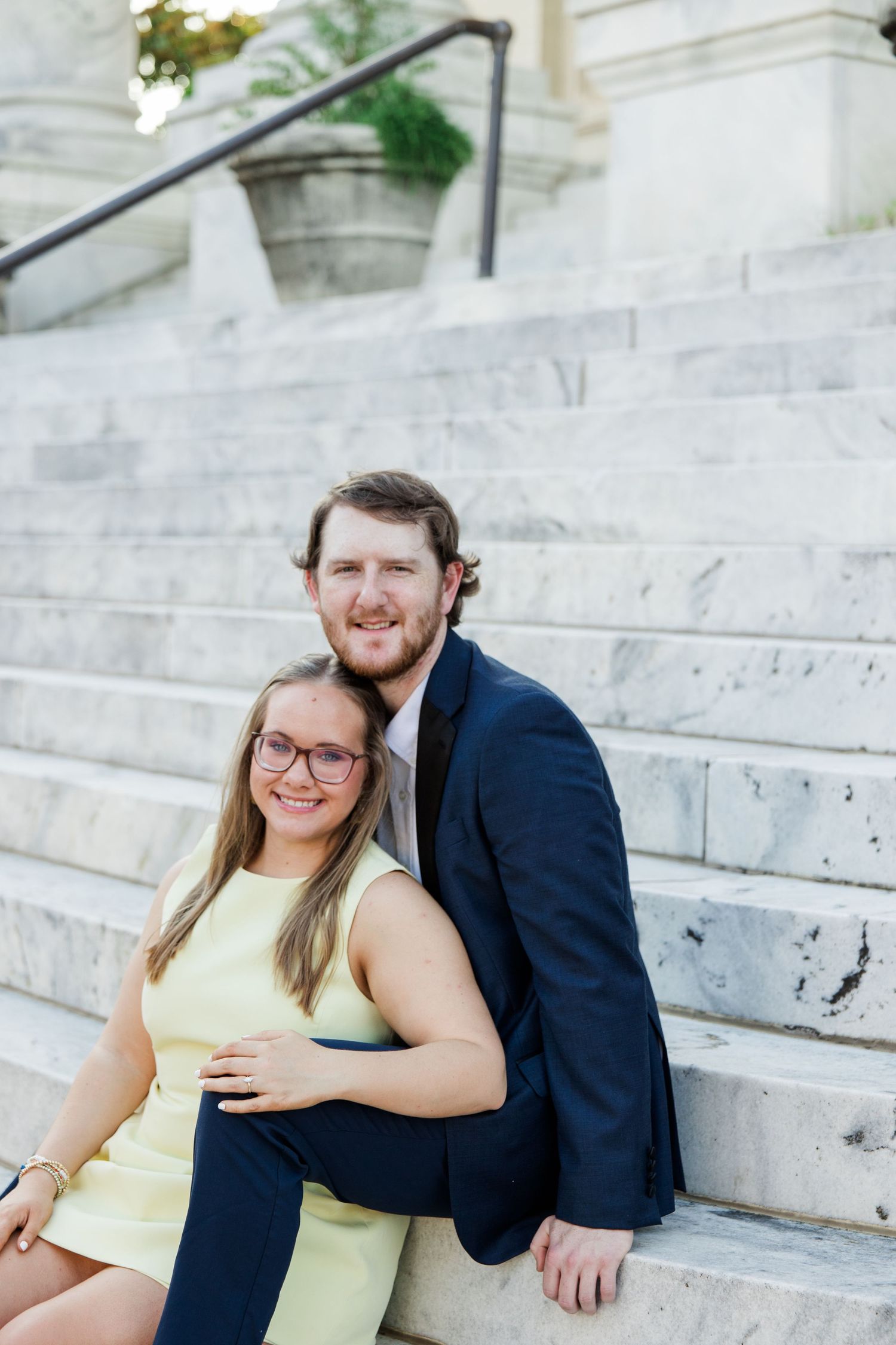 A smiling couple in formal attire sits on marble steps, woman in yellow dress with glasses, man in navy suit embracing her.