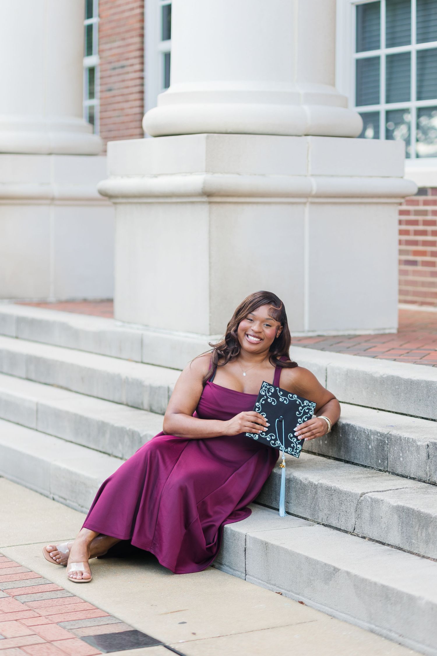 Smiling graduate in purple dress sits on steps holding graduation cap at brick university building.