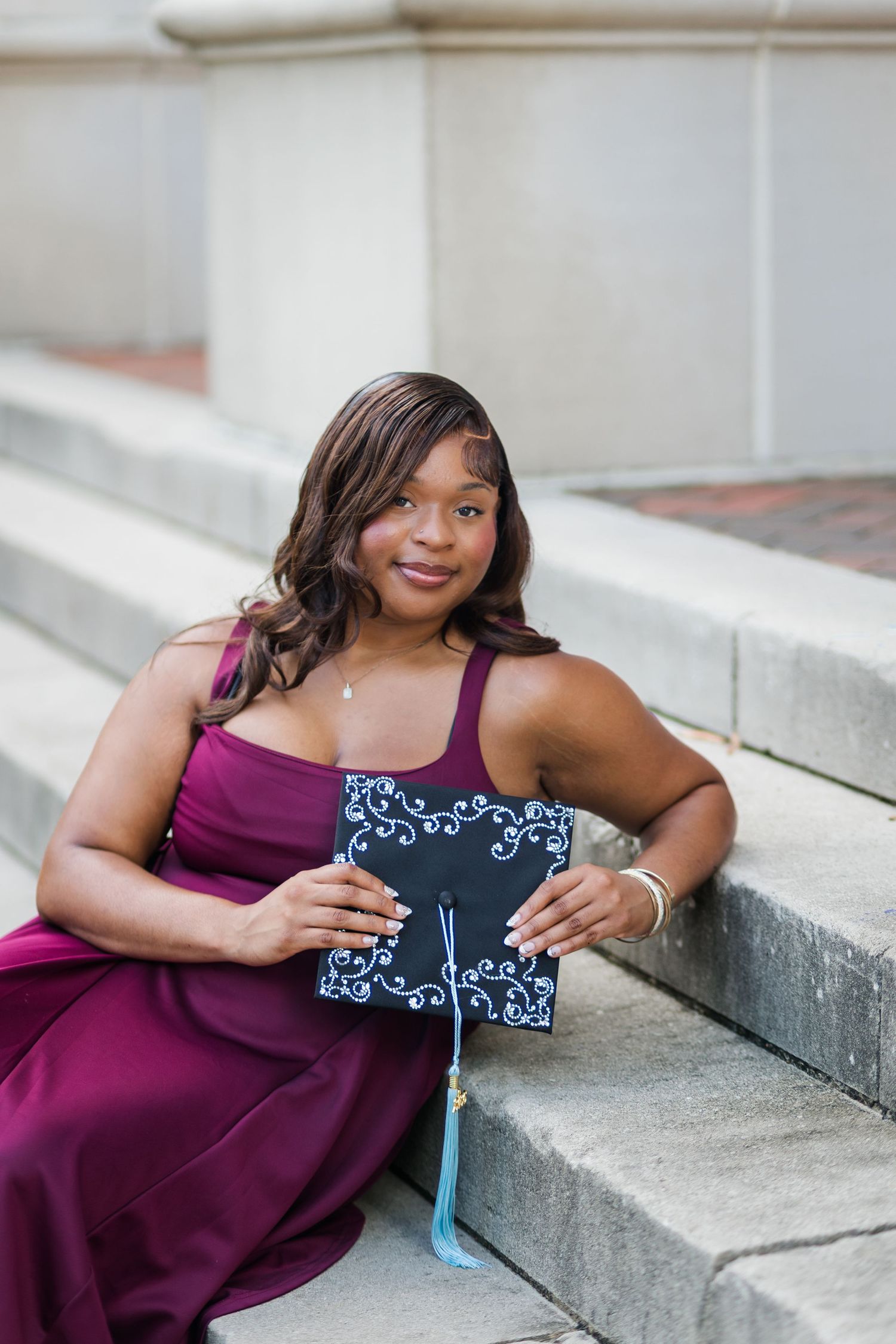 A young woman in a burgundy gown sits on steps holding a decorated graduation cap with blue tassels.