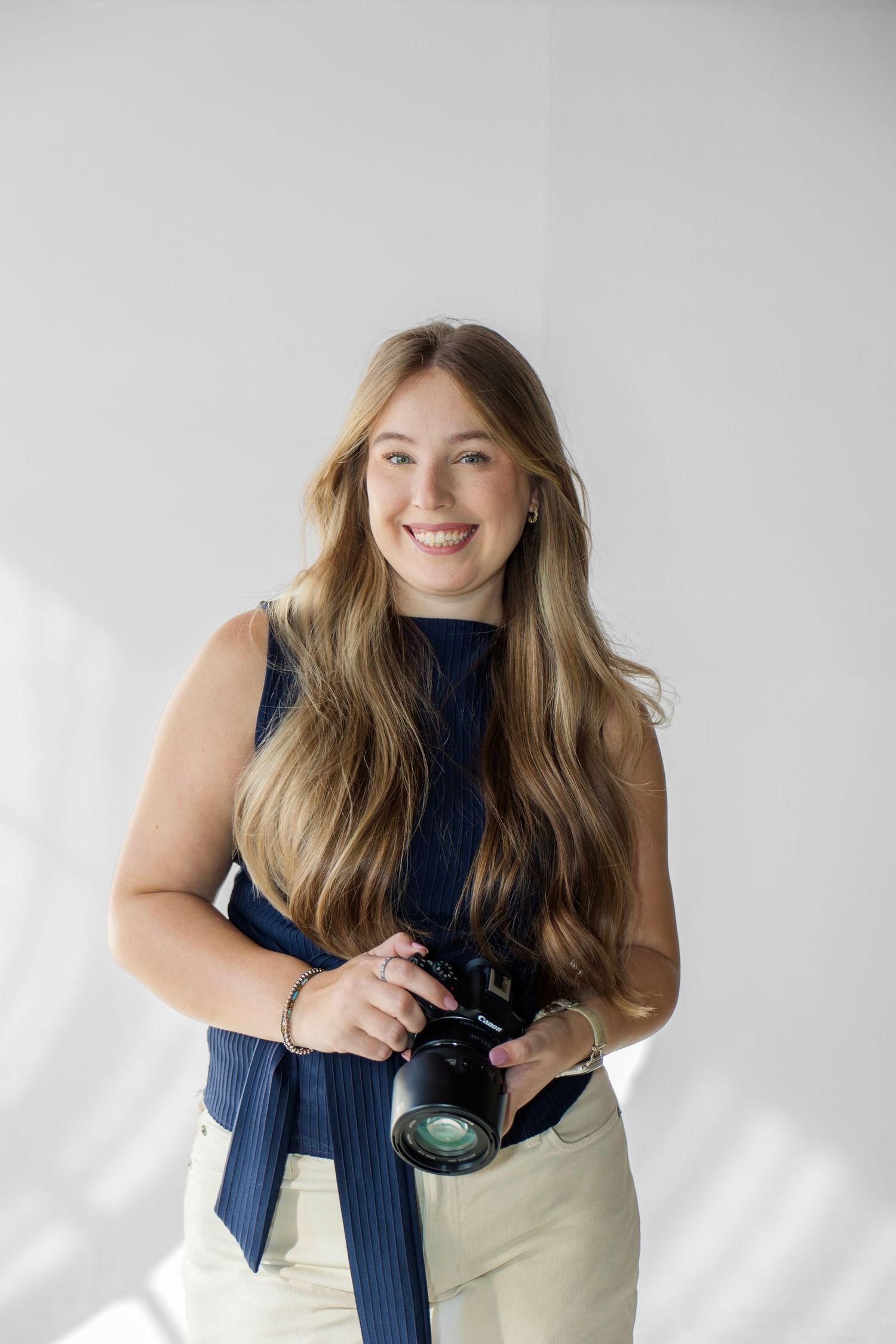 Smiling young woman with long blonde hair holding a camera, wearing a navy top and beige pants against a white background.