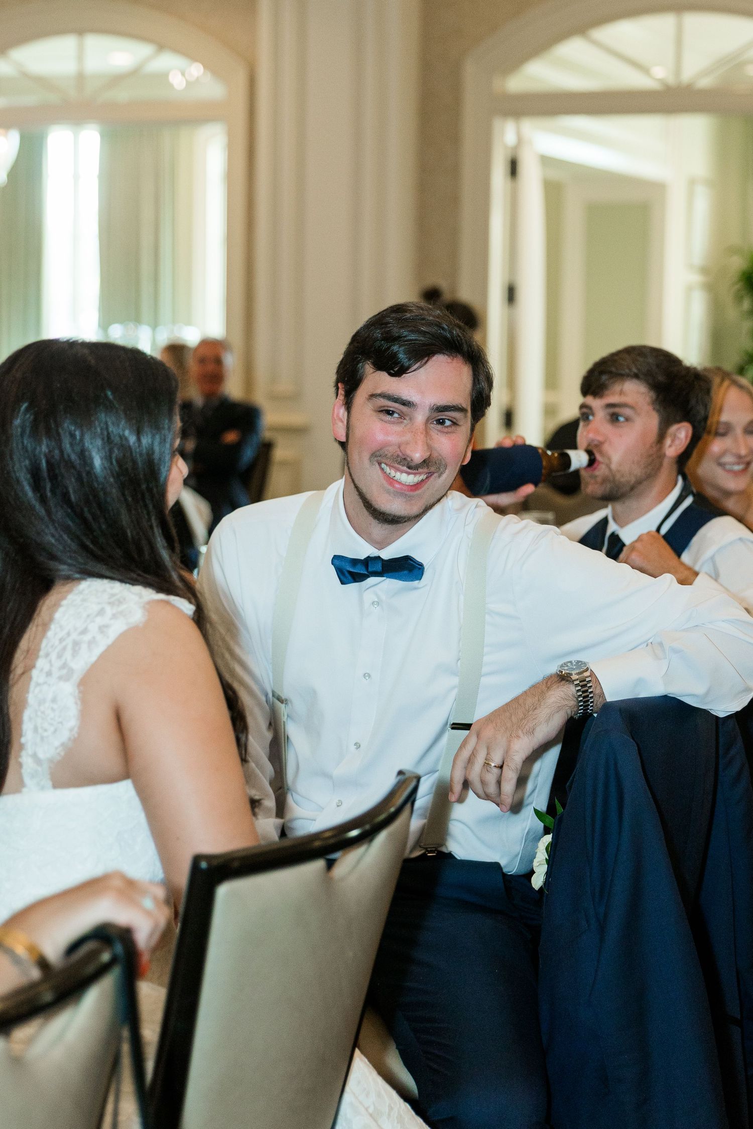 Smiling groom in white shirt and blue bow tie chatting with bride at wedding reception.