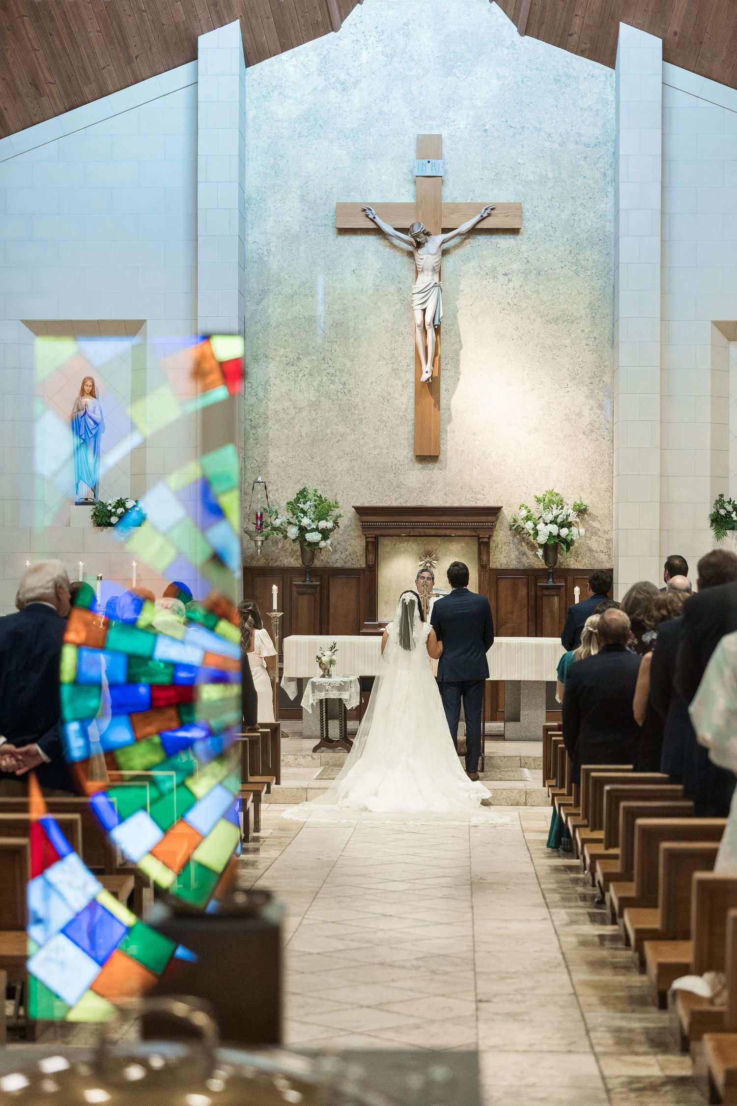 Bride and groom stand at altar during Catholic church wedding ceremony with crucifix and stained glass.