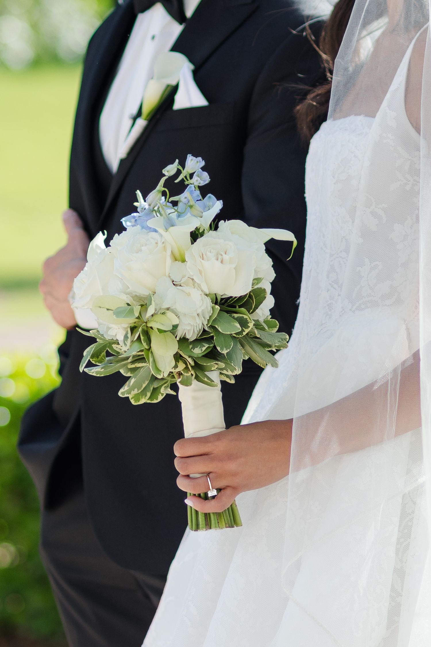 Bride in white lace gown holding elegant bouquet of white roses and blue flowers beside groom in black tuxedo.