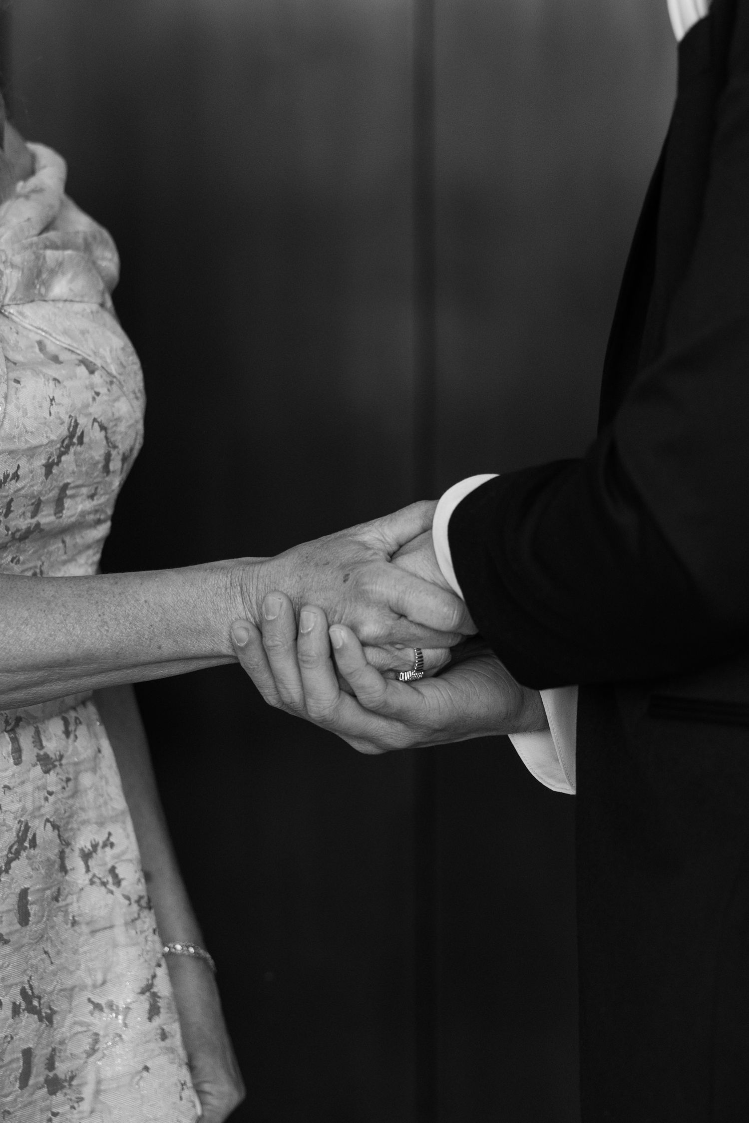 Black and white photo of couple holding hands, wearing wedding attire, with a diamond ring visible.