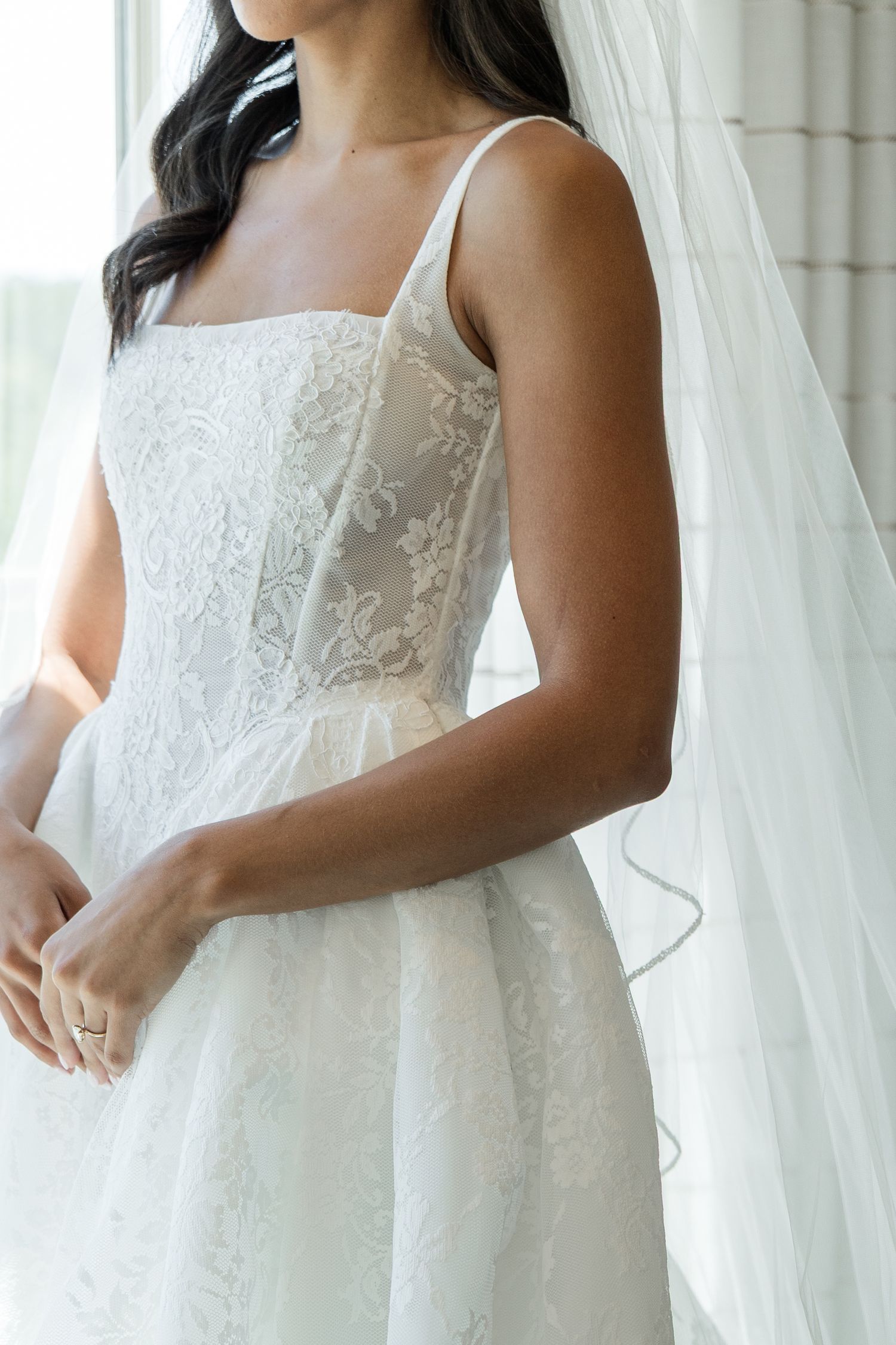 Bride in a lace square-neck wedding gown with sheer corset bodice and flowing veil, standing by a window.