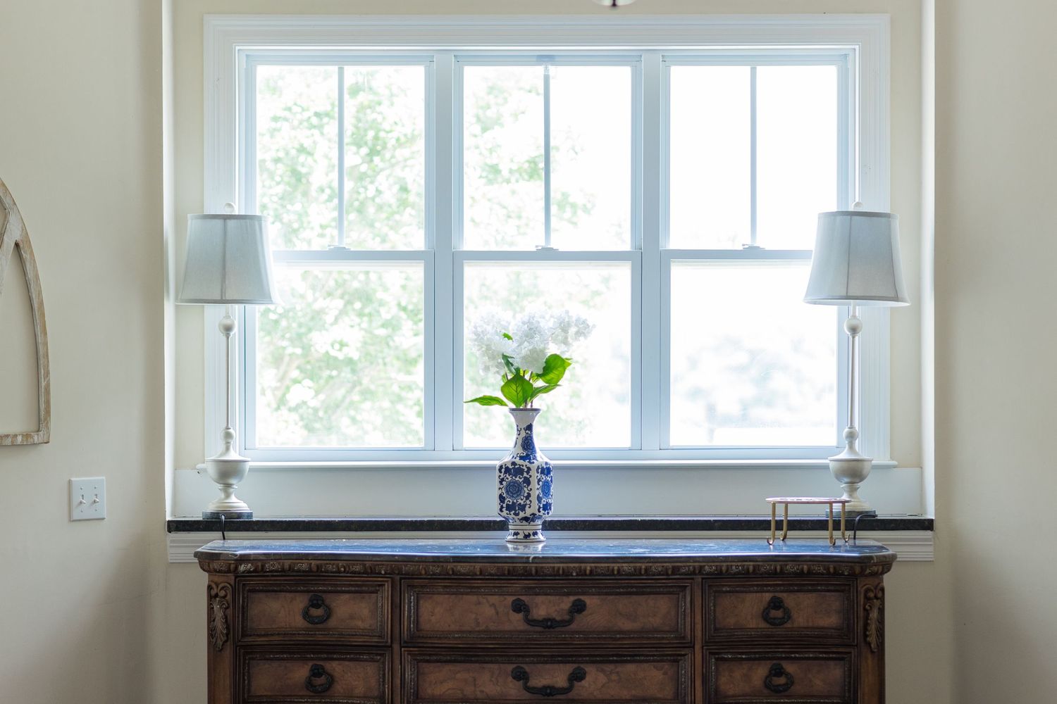 Antique wooden dresser with blue and white floral vase and twin lamps beneath a bright double-hung window.