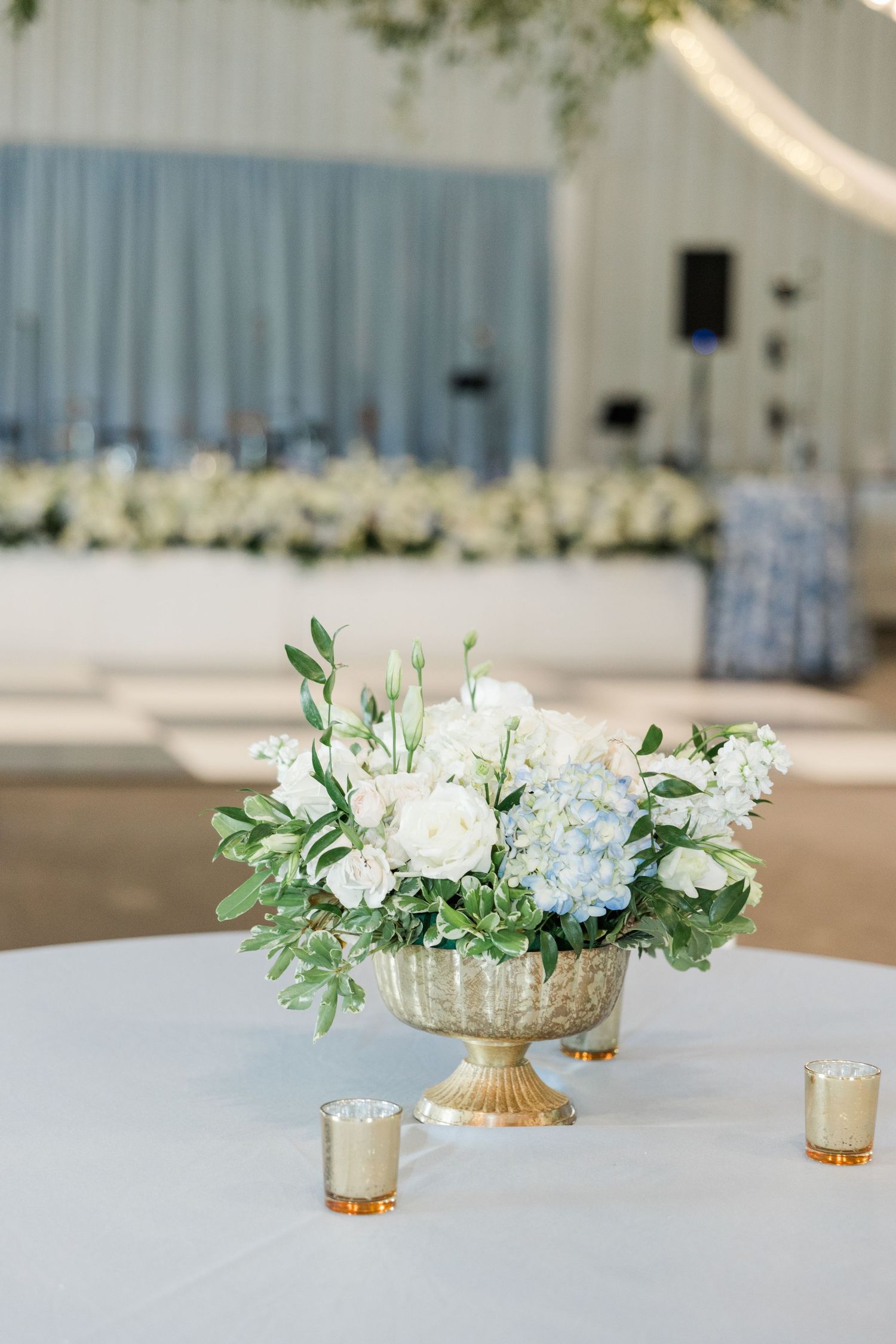 Elegant gold urn centerpiece filled with white roses and blue hydrangeas on white tablecloth at wedding reception.