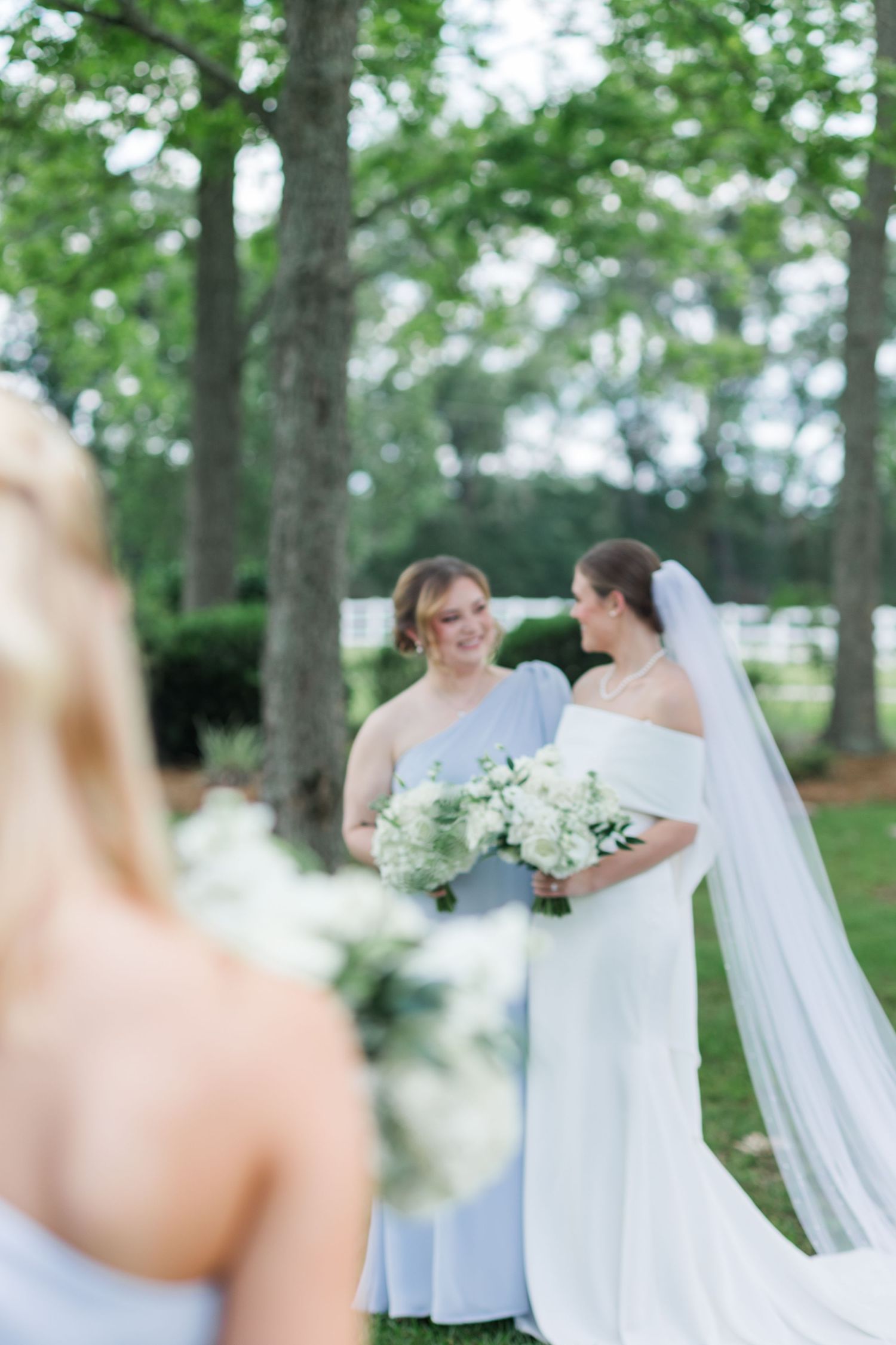Bride in off-shoulder gown and veil holding white bouquet with bridesmaid smiling outdoors.