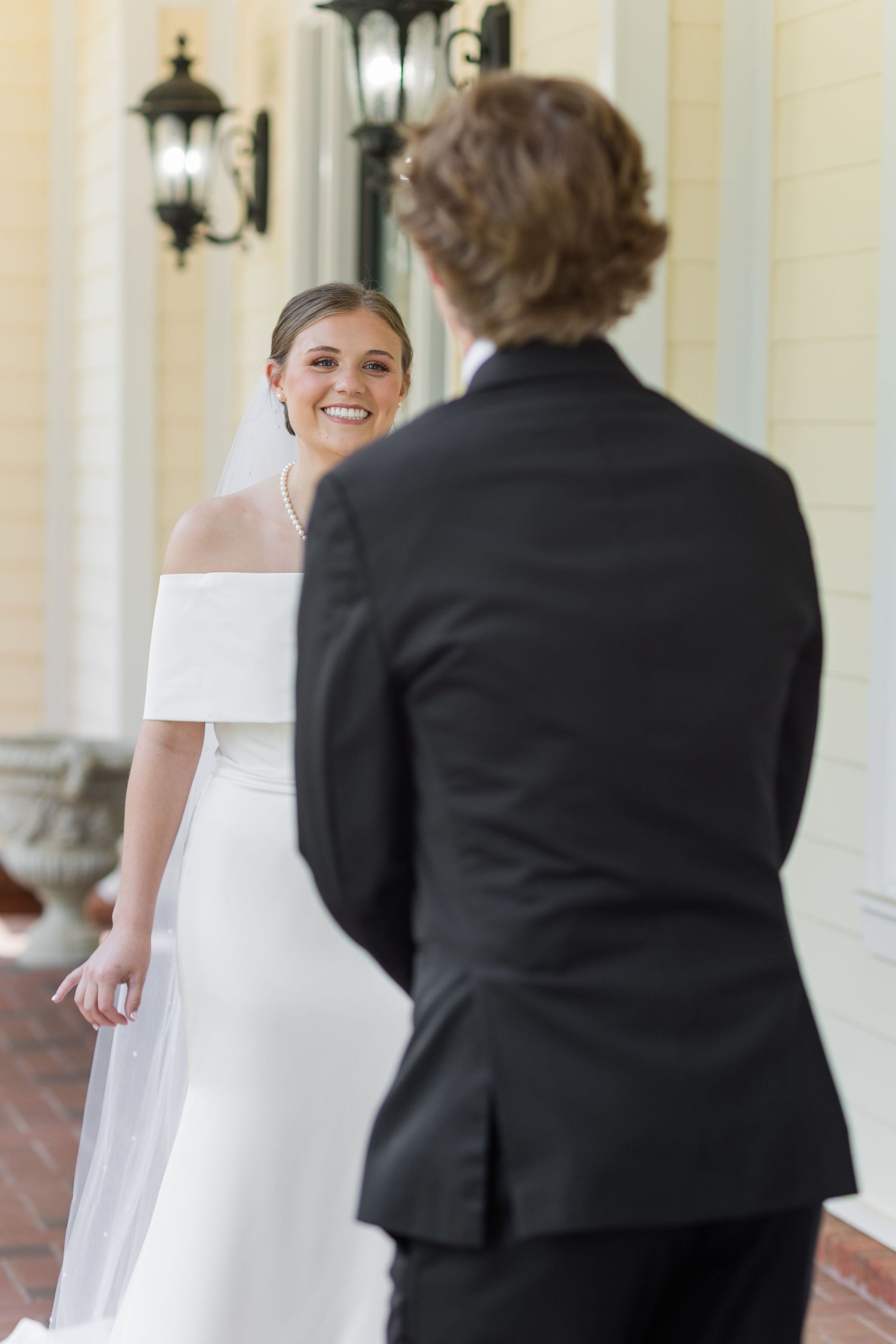 Bride in off-shoulder white gown smiling at groom during first look moment on venue porch.