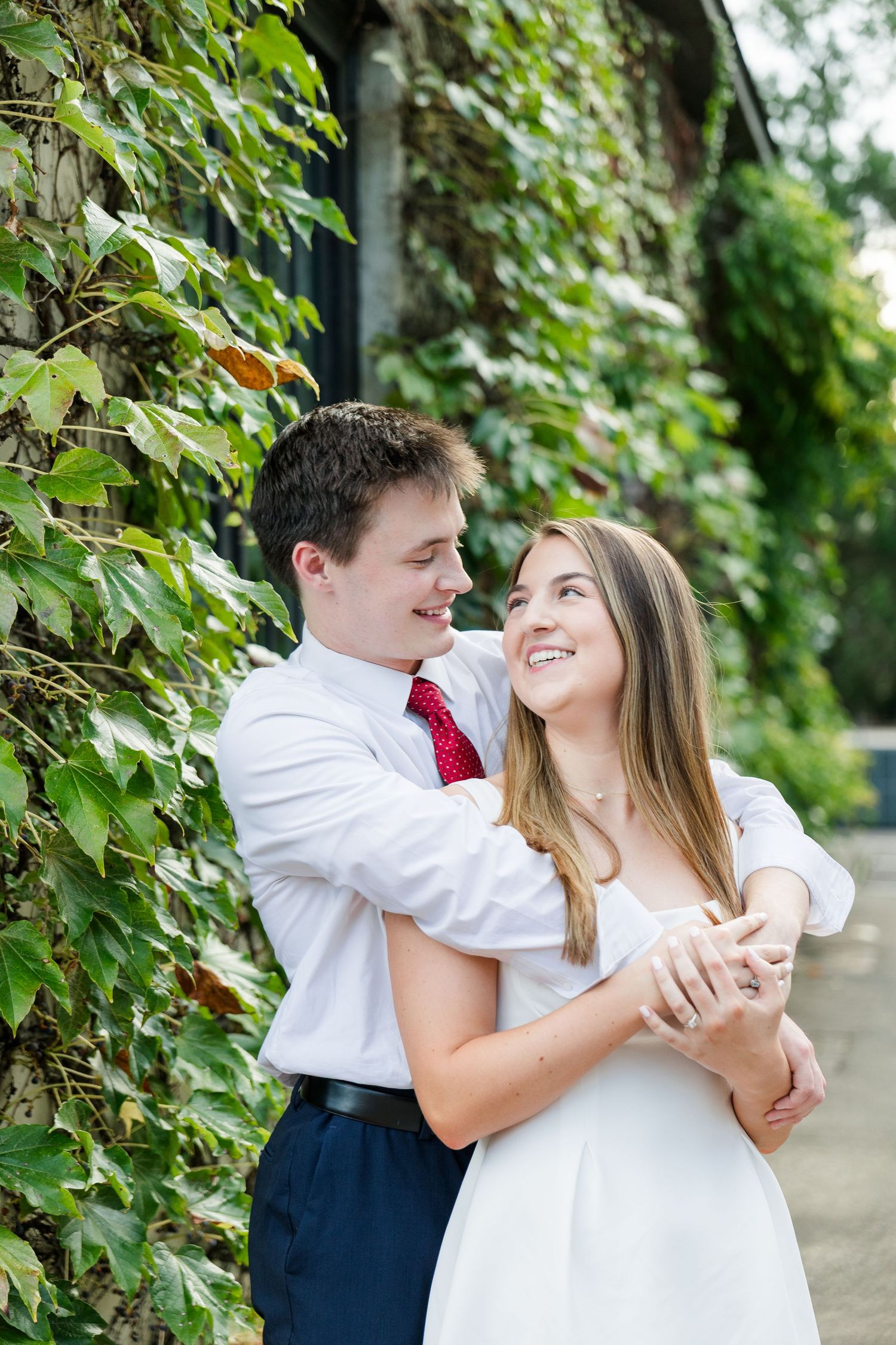 Happy couple hugging and laughing against ivy-covered stone wall, woman in white dress, man in red tie.