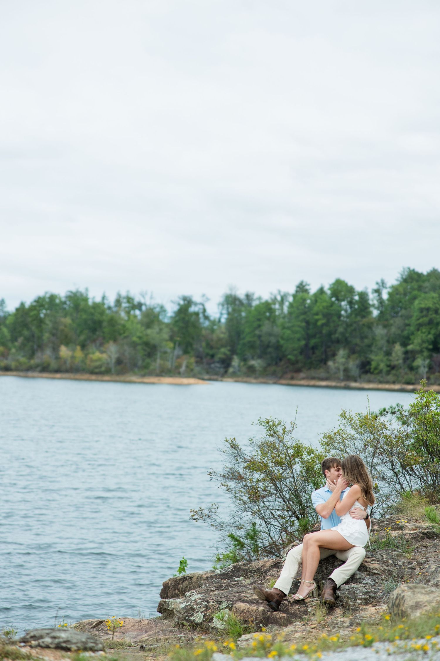 Couple sits close together on a rocky outcrop overlooking a calm lake with a forested shoreline under a soft overcast sky.