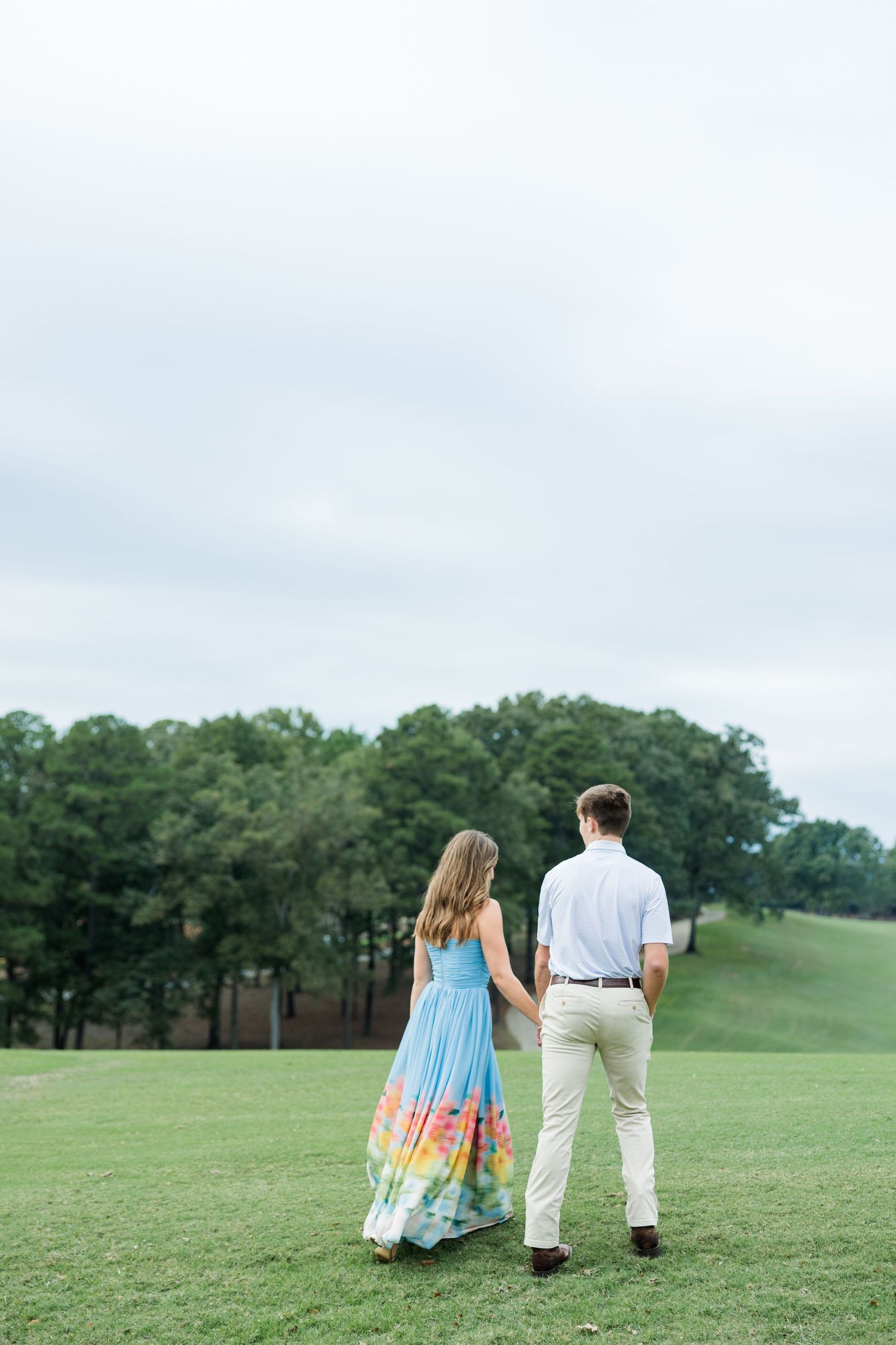 Couple holds hands and walks away across a grassy hill toward distant trees, her colorful watercolor-hem gown flowing in the breeze.
