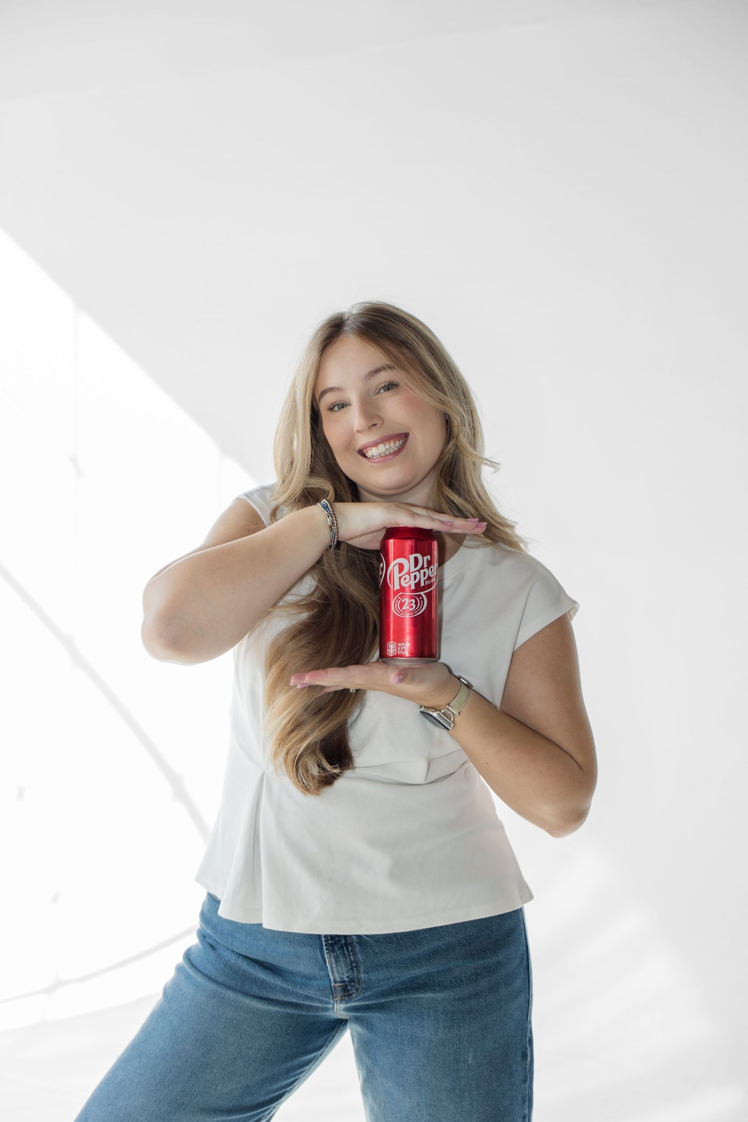 Smiling young woman holding a red Dr Pepper can with both hands against a white background.