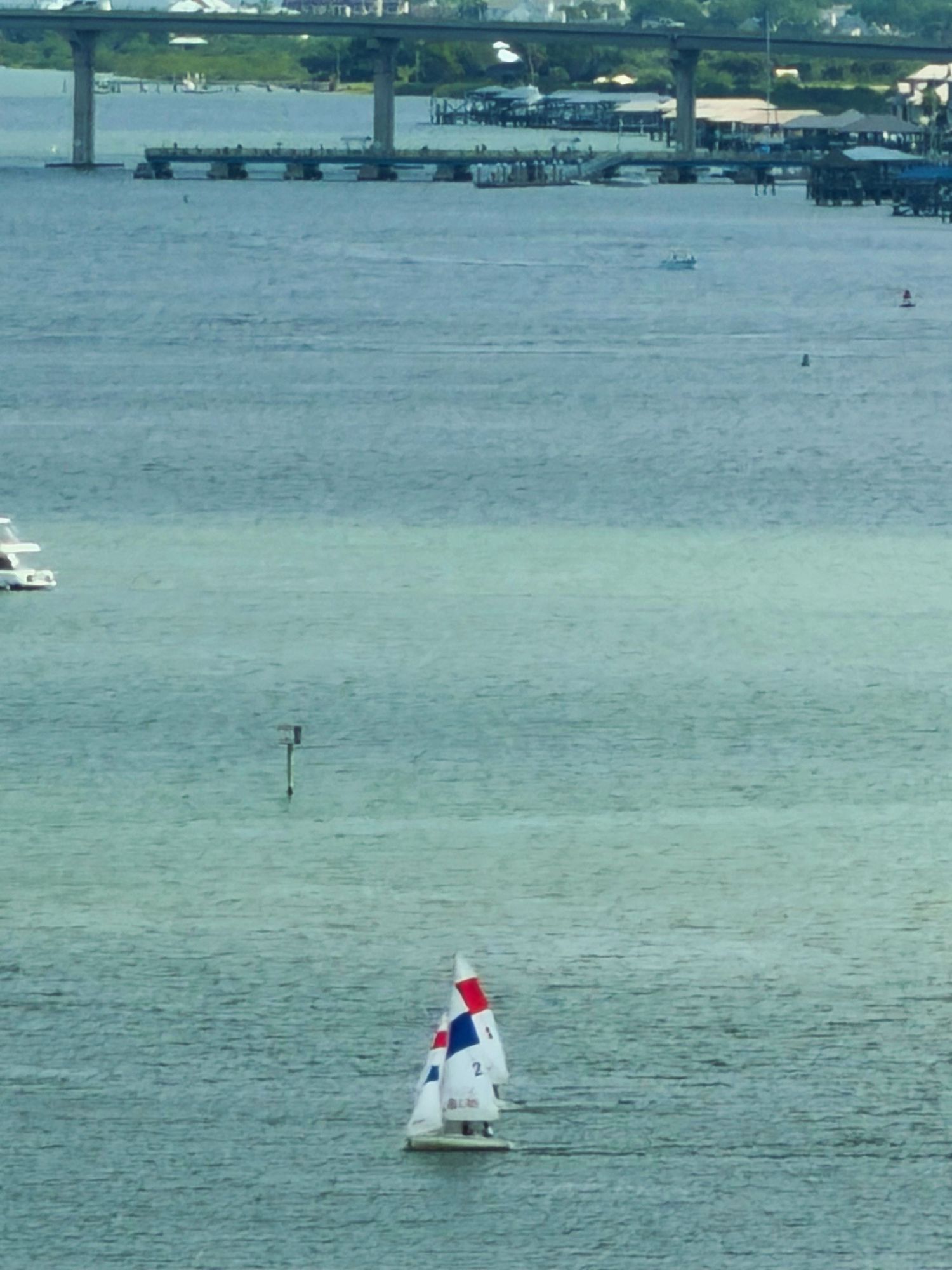 Sailboat with red and white sails gliding on calm blue water near a bridge.