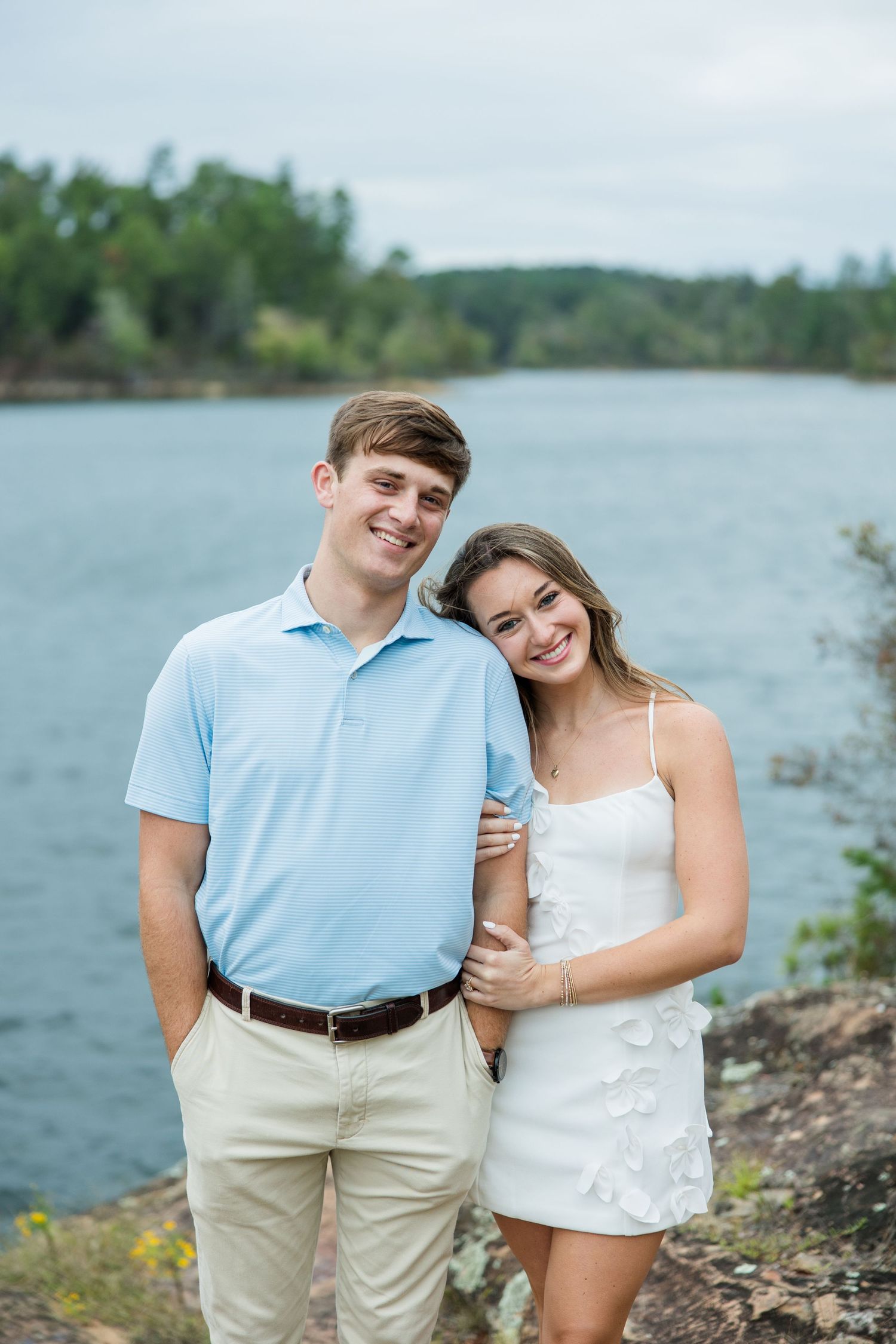 A smiling couple in light summer outfits poses together on a rocky shoreline with a calm lake and tree-lined banks behind them.