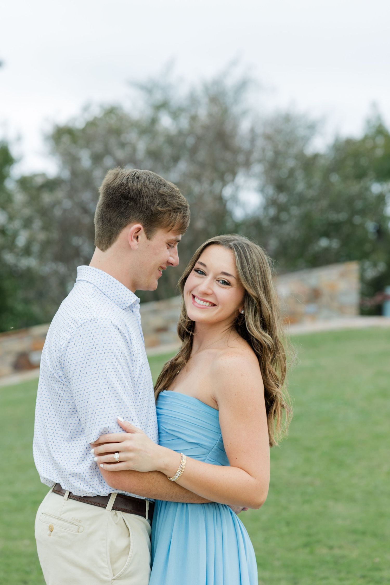 Smiling couple embracing during an outdoor engagement photo session on a grassy lawn, she in a blue dress and he in a white shirt.