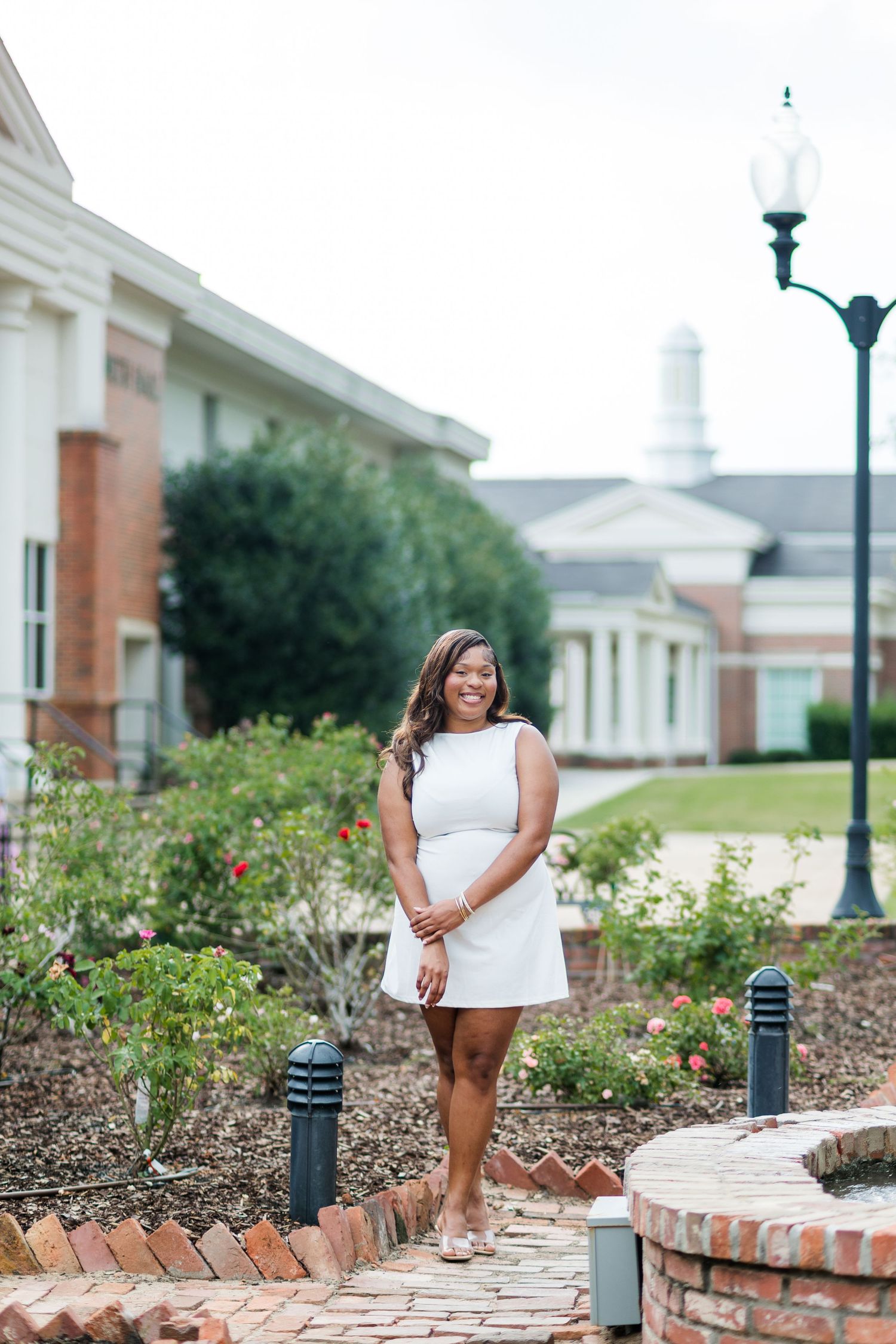Young woman in white dress posing on brick path in front of elegant campus building with lush gardens.