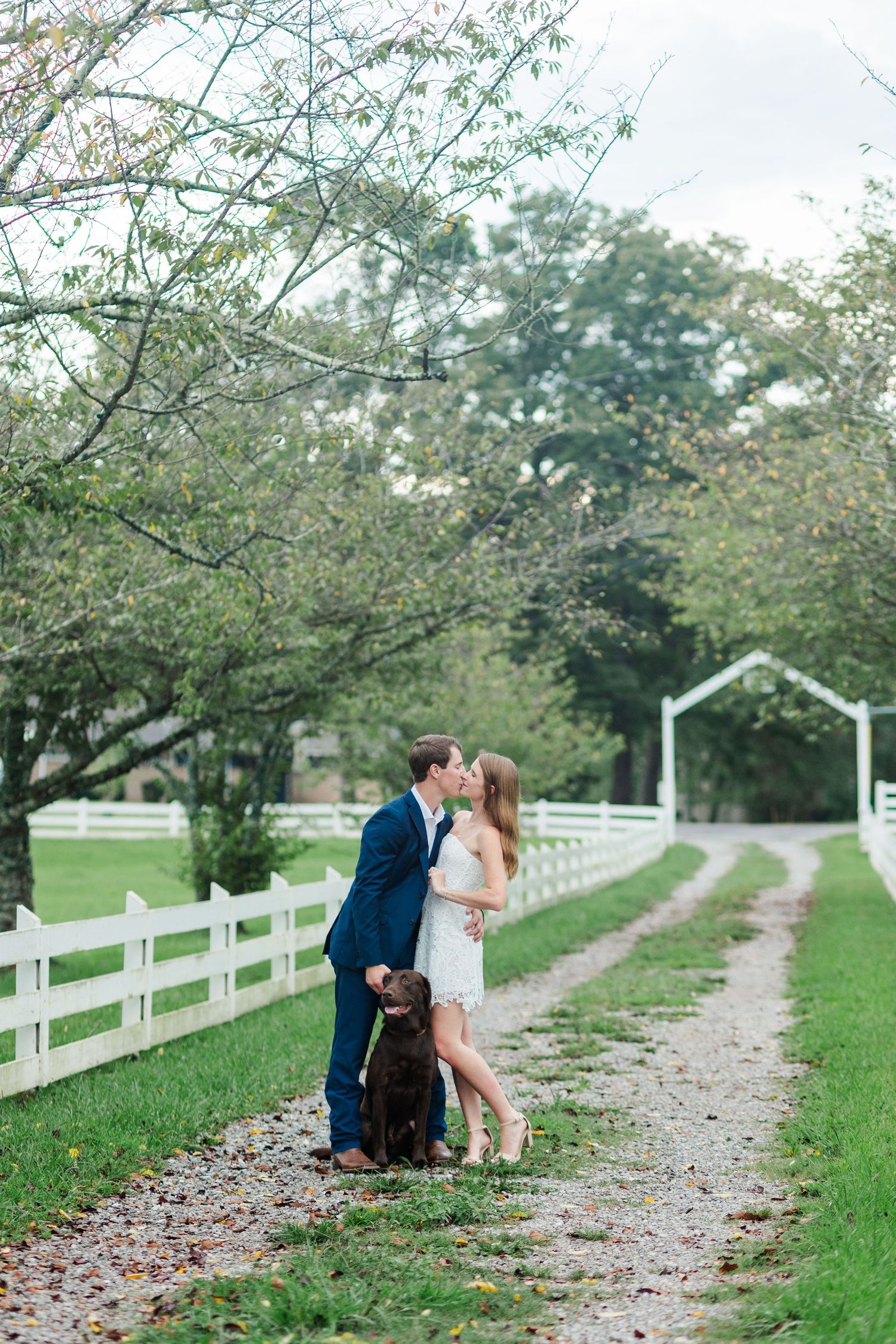 Couple kissing on a tree-lined gravel path with white fence and brown dog during outdoor engagement photo session.