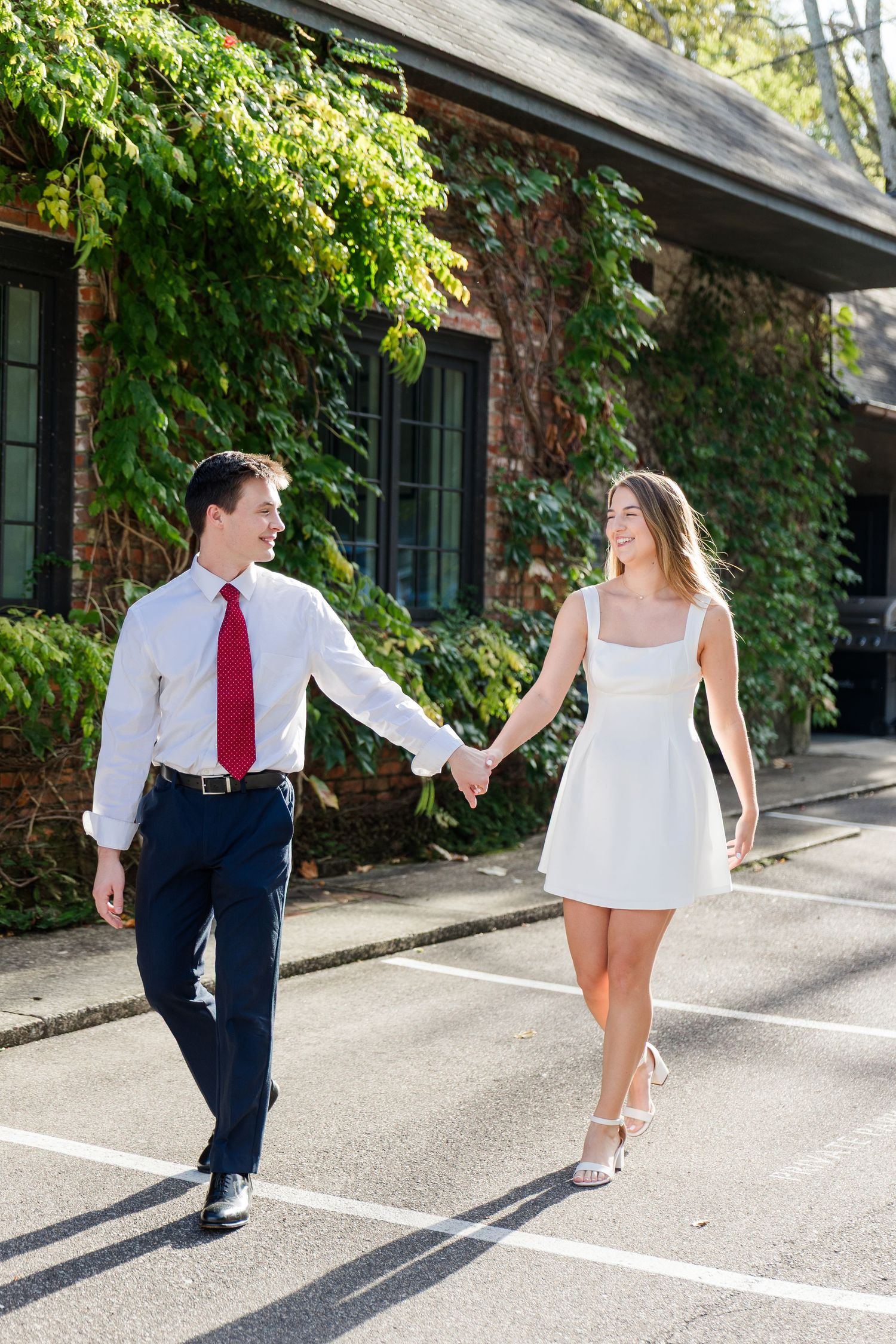 A couple holds hands walking in a parking lot, man in white shirt and red tie, woman in white mini dress and heels.
