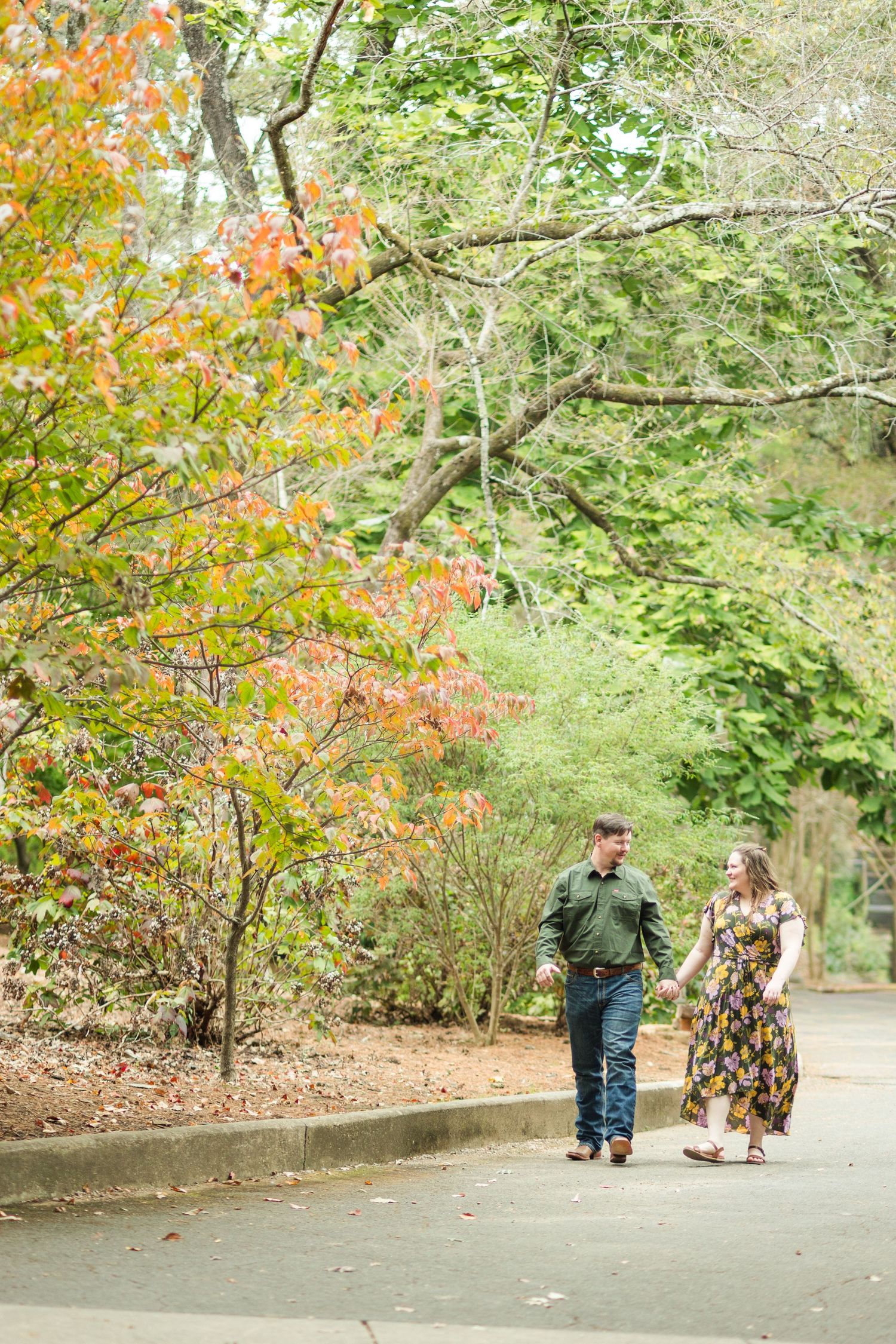 Couple walking hand in hand on a tree-lined path surrounded by colorful autumn foliage in a lush park.