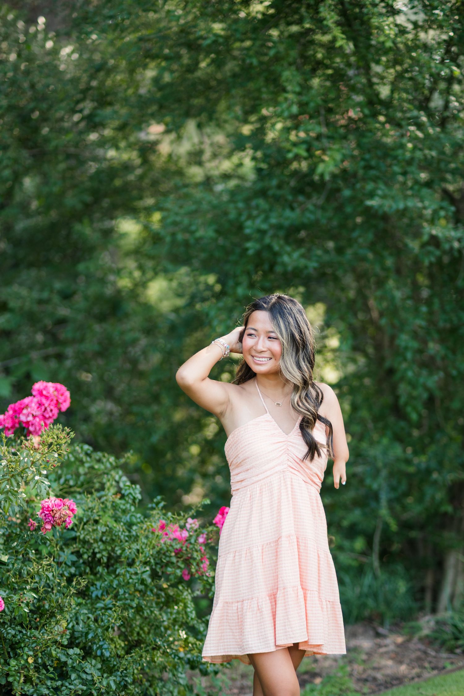 A smiling young woman in a peach sundress poses beside vibrant pink roses in a green garden.