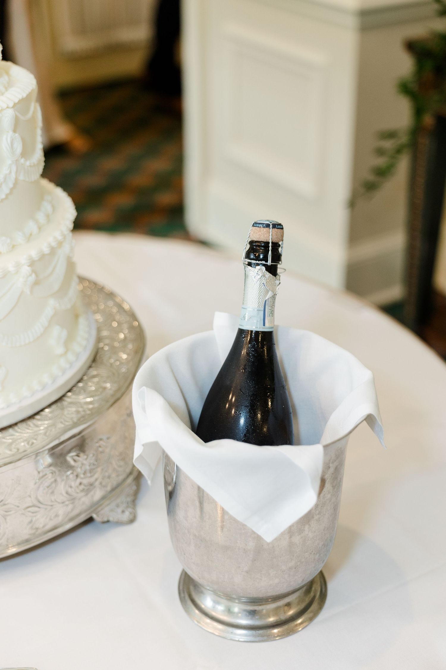 Champagne bottle chilling in silver ice bucket beside a white tiered wedding cake on a white tablecloth.