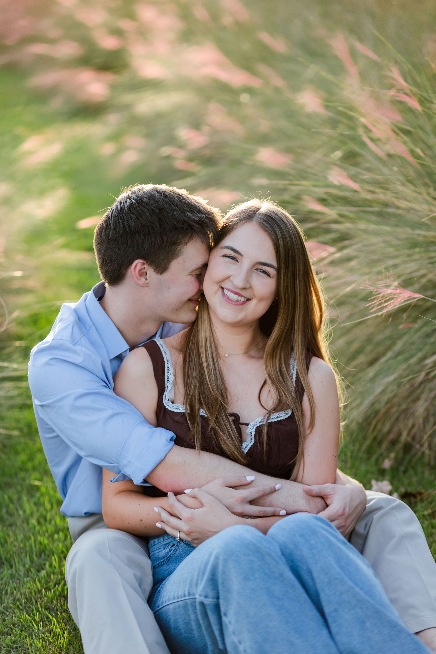 Happy couple sitting in grass, woman smiling as man whispers in her ear near ornamental grasses.