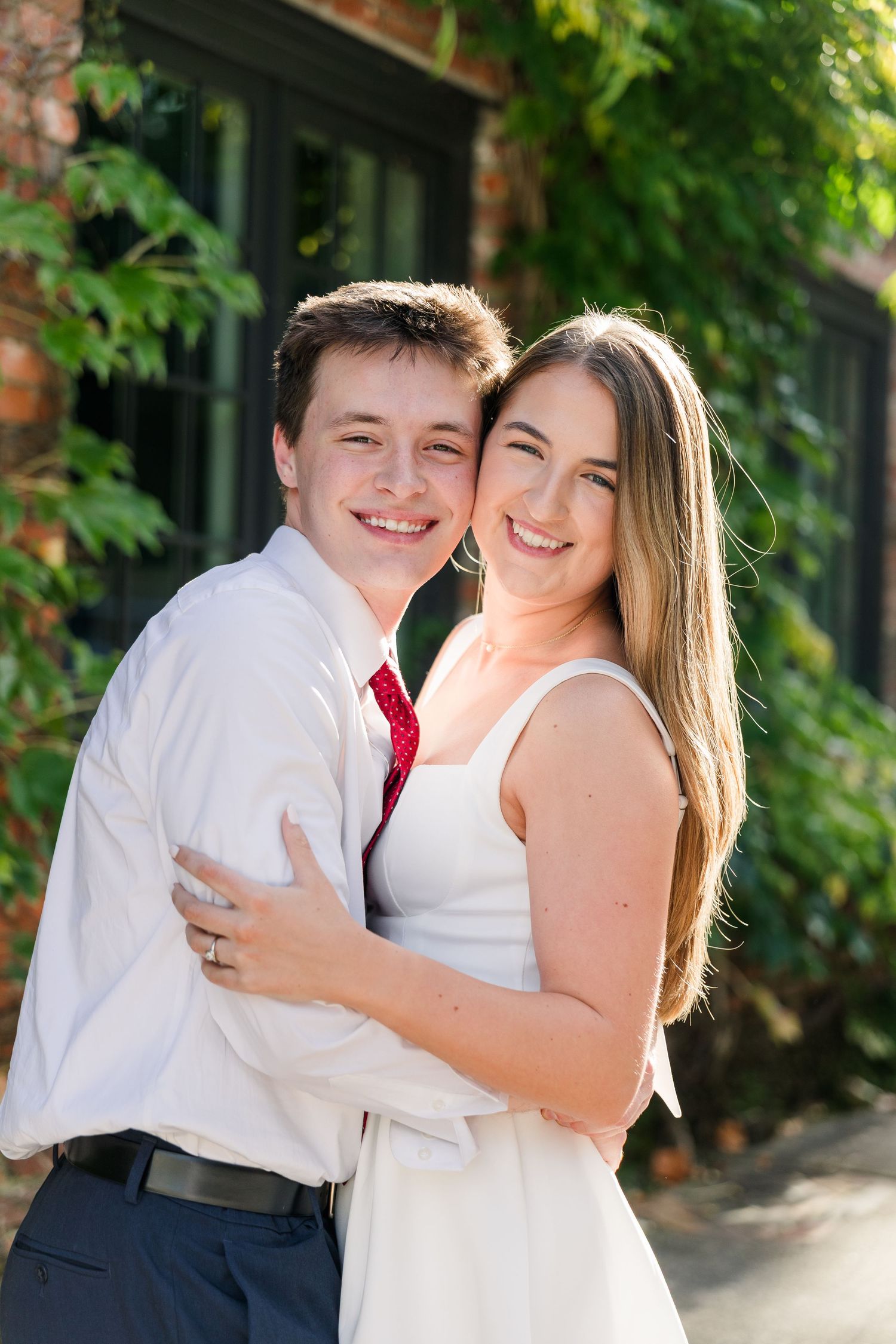 A smiling young couple embraces outdoors near a brick building covered in green ivy.