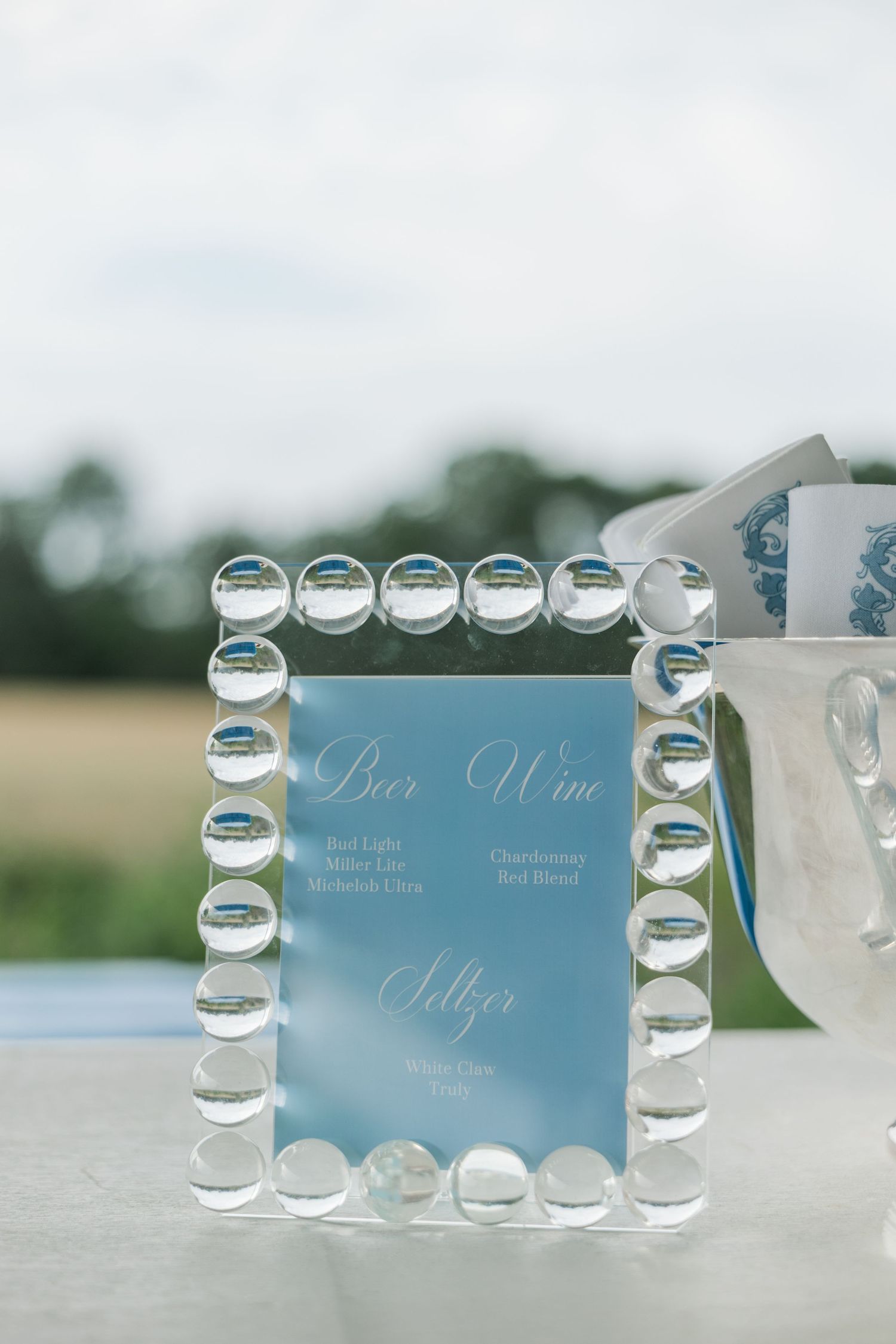 A crystal-framed bar menu sign listing beer, wine, and seltzer options at an outdoor wedding reception.