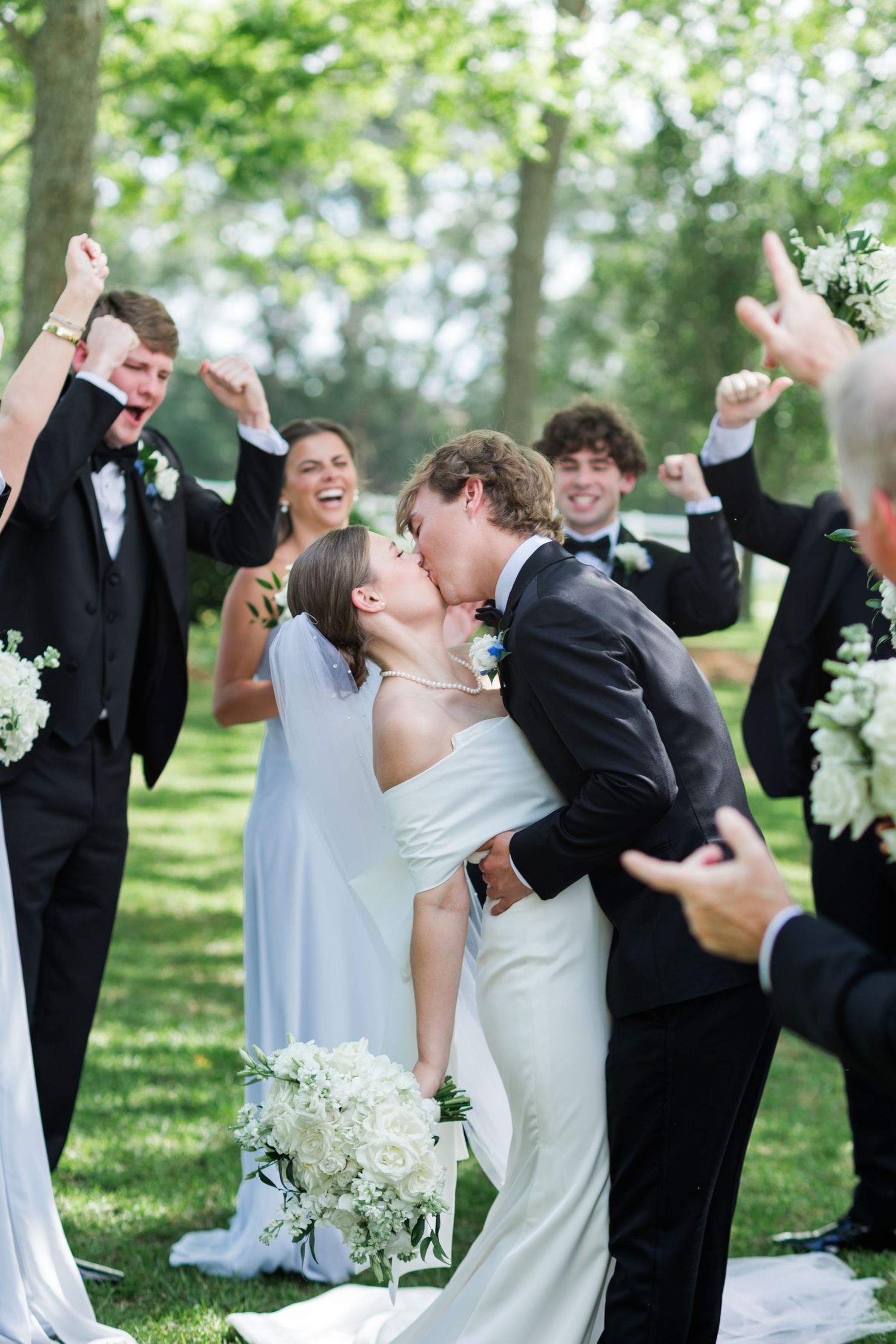 Bride and groom share a kiss surrounded by cheering wedding party in elegant black and white attire outdoors.