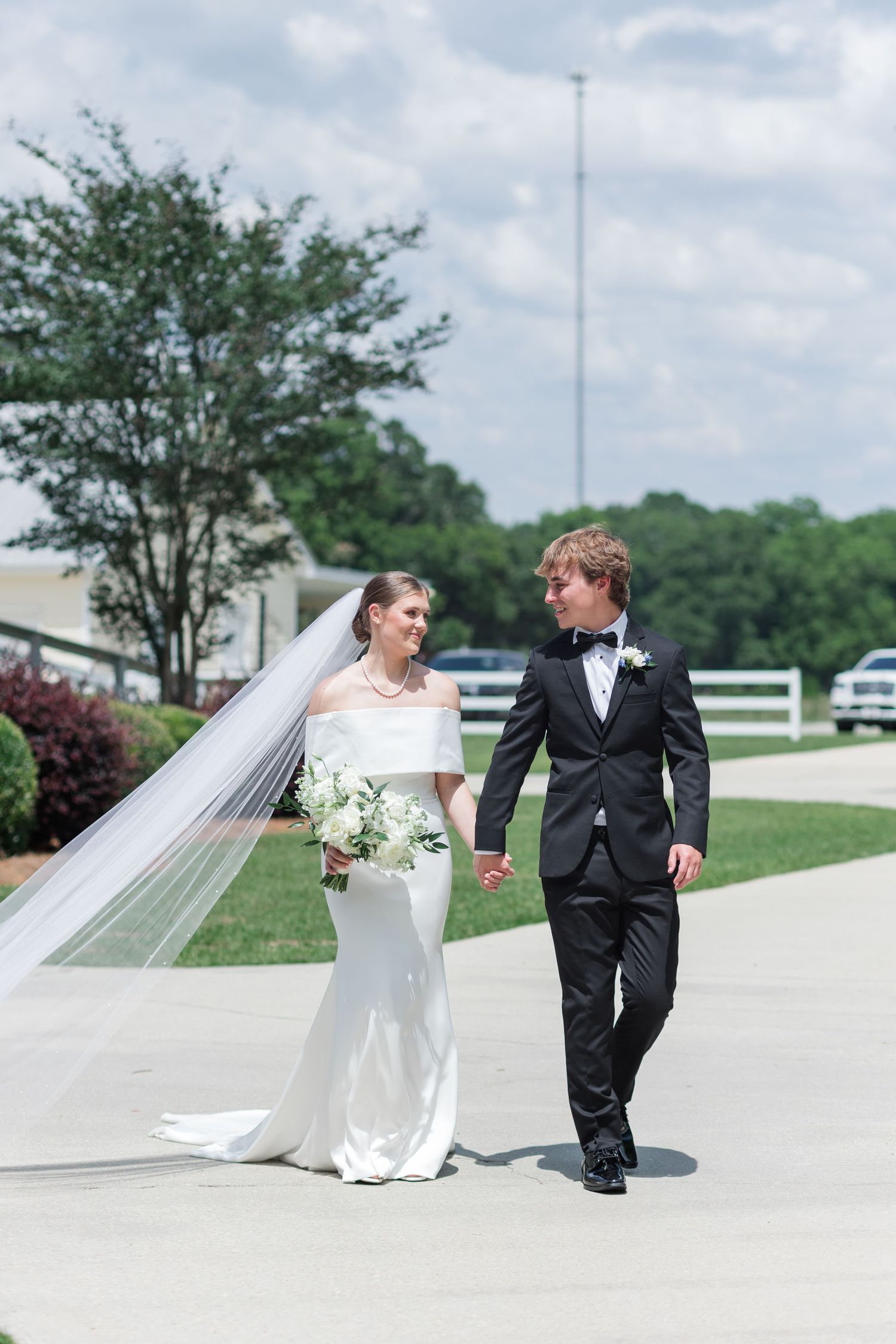 Bride and groom walking hand-in-hand outdoors, bride wearing flowing veil and holding white bouquet.