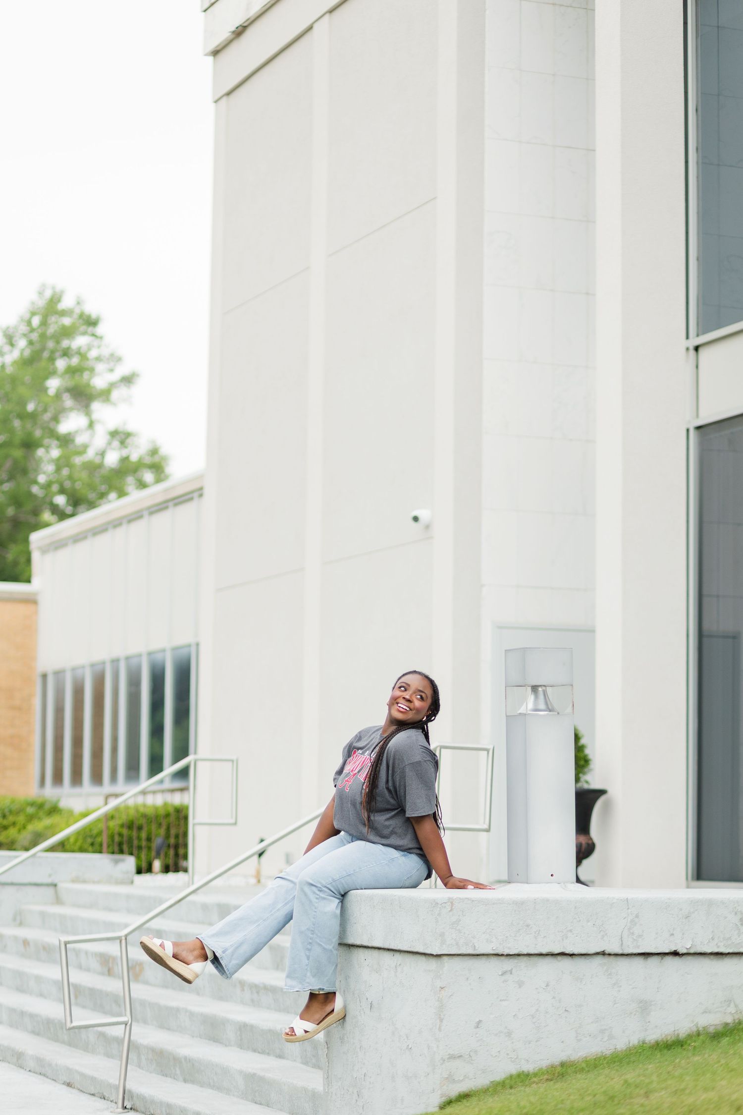 Young Black woman in gray shirt and jeans sitting on white concrete steps outside modern building.