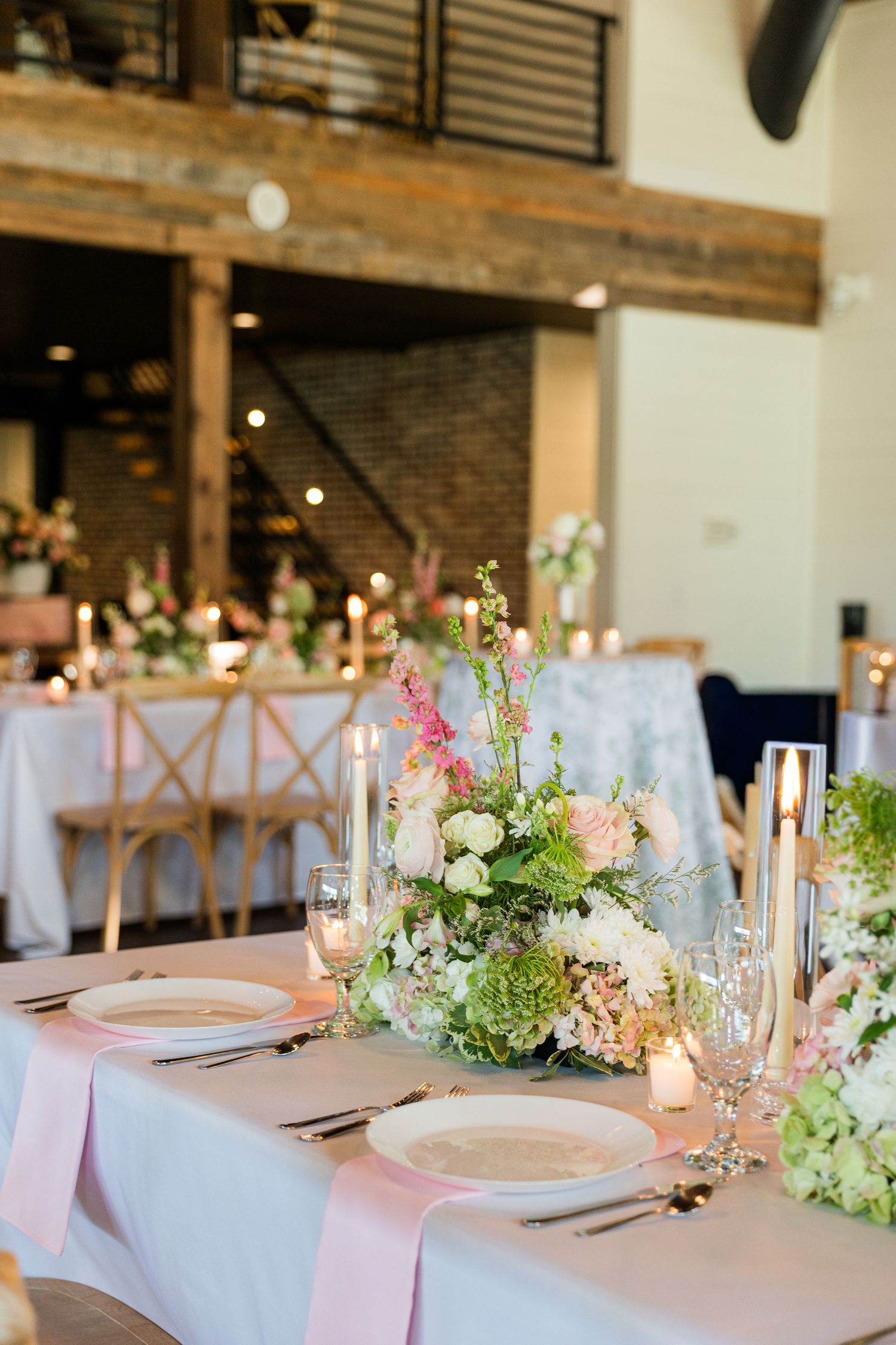 Elegant wedding reception table with pink linens, floral centerpieces of roses and greenery, candles, and rustic barn venue backdrop.