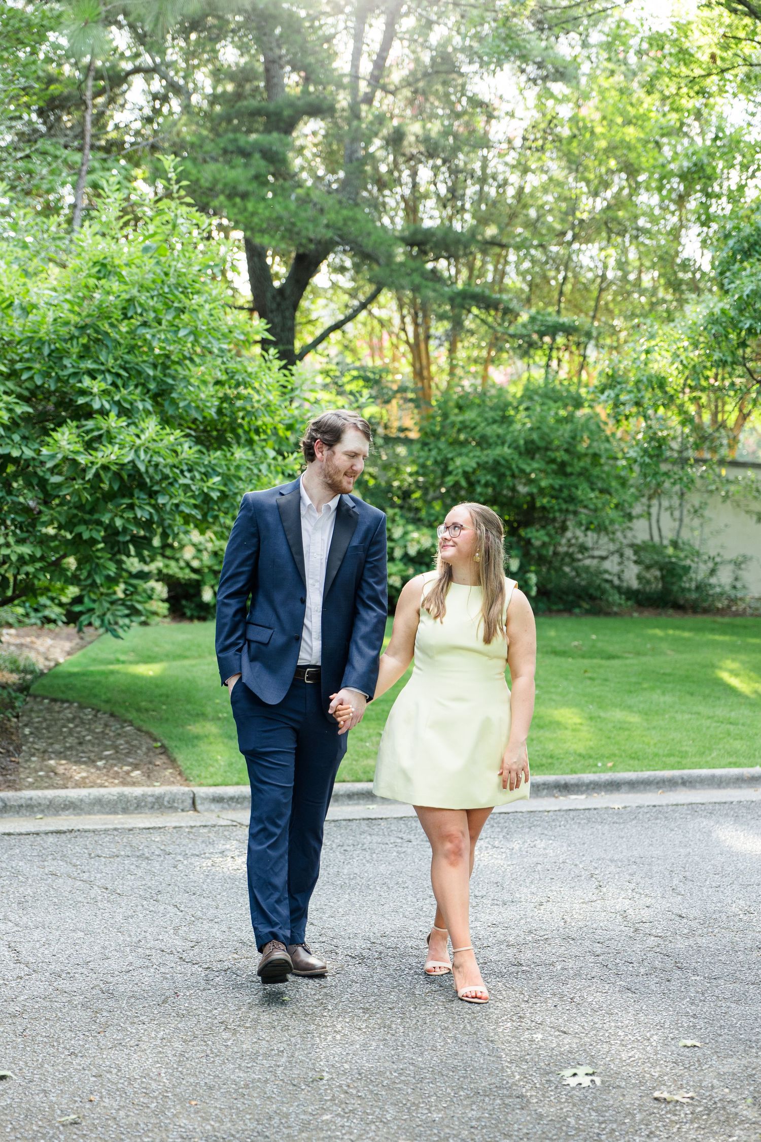 Couple holding hands and smiling while walking outdoors in a lush green garden, man in navy suit, woman in yellow dress.