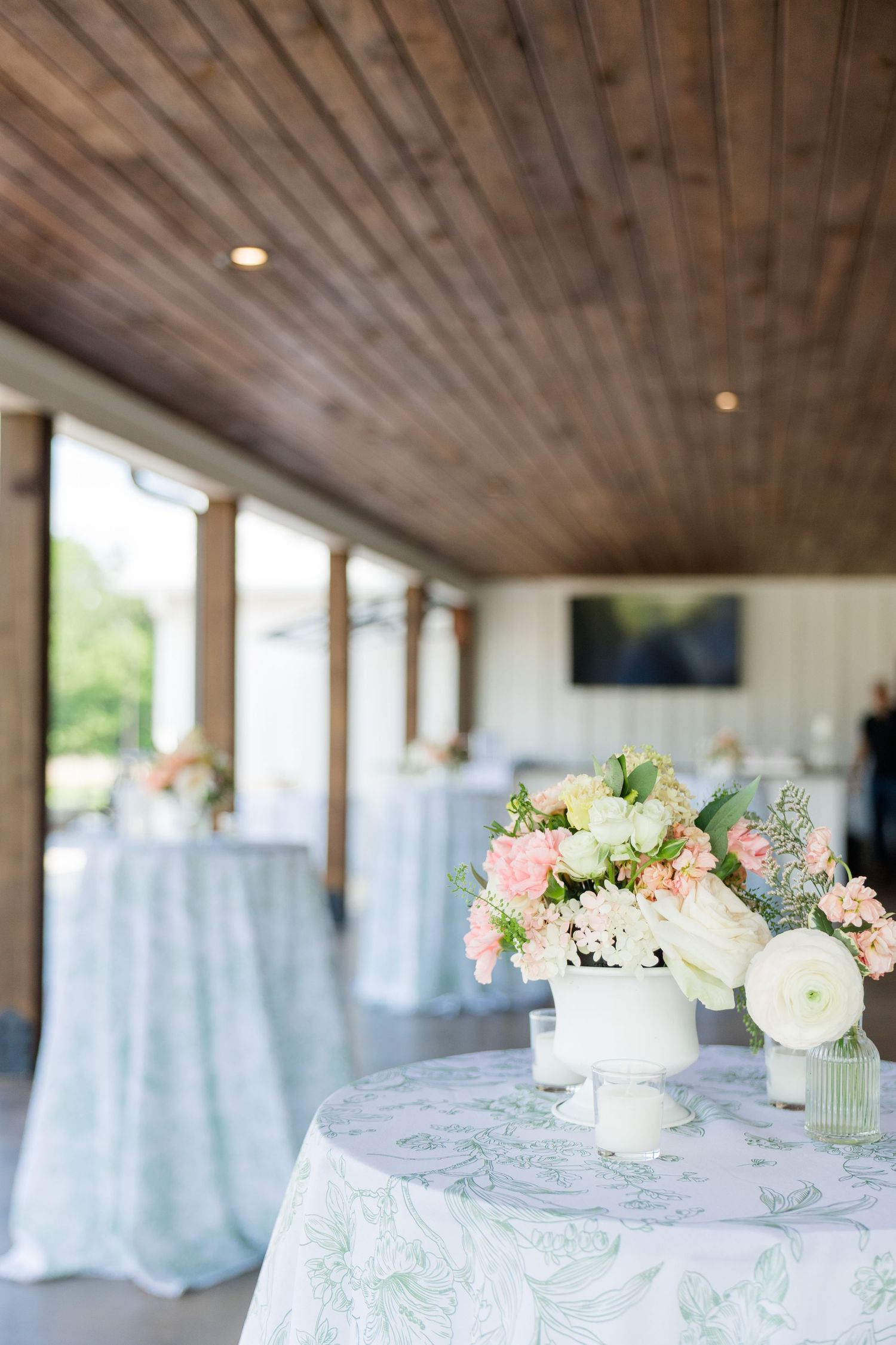 Elegant wedding reception with floral centerpiece of pink and white blooms on a patterned tablecloth under a wood ceiling.