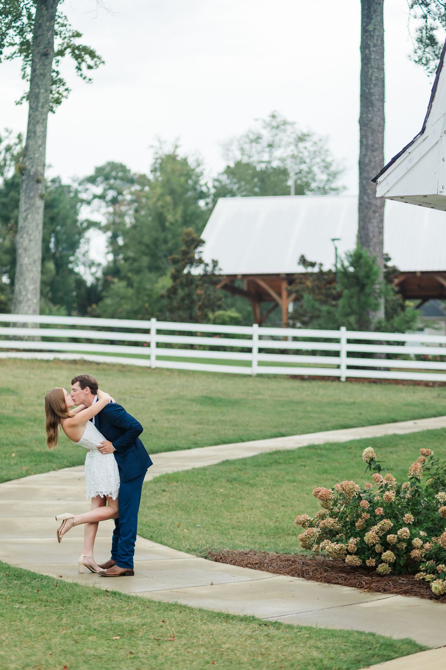Groom lifting bride for a romantic kiss on a garden pathway with white fence and barn in background.