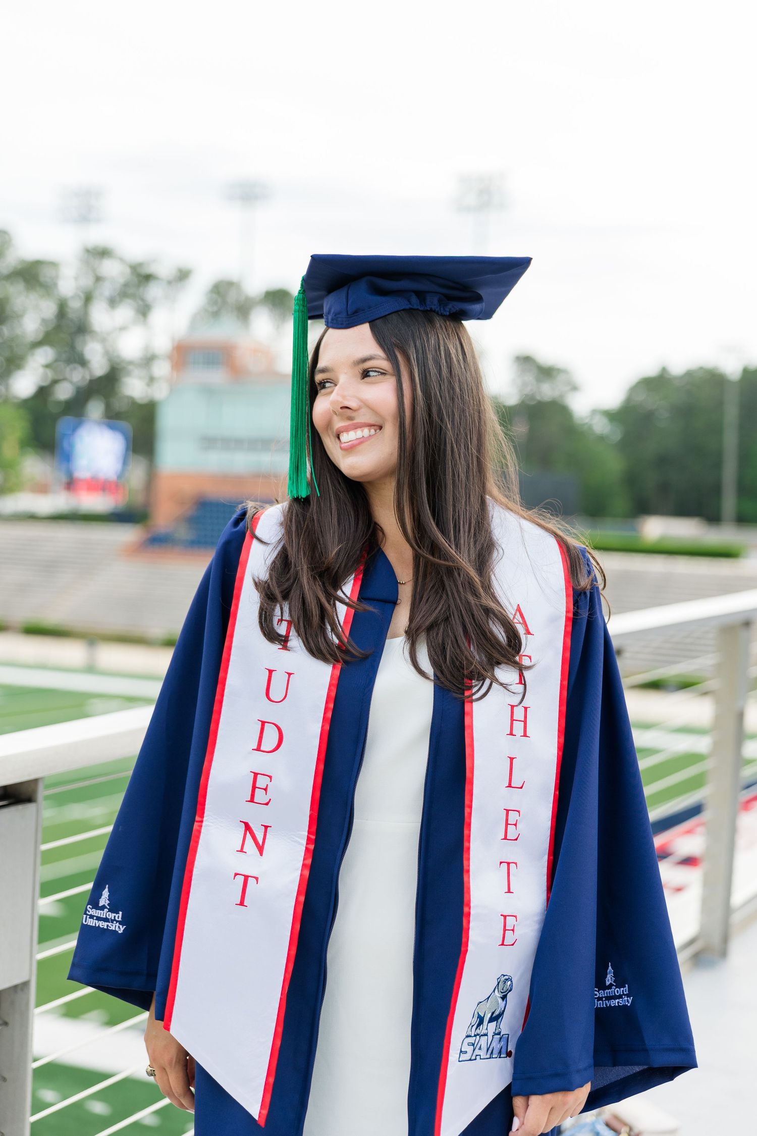 Smiling female graduate in navy cap and gown with Student Athlete stole at university stadium.