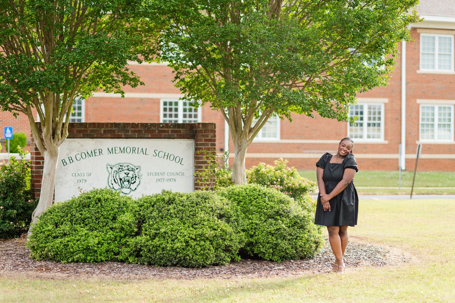 Young woman in black dress poses by B.B. Comer Memorial School brick sign surrounded by trees.