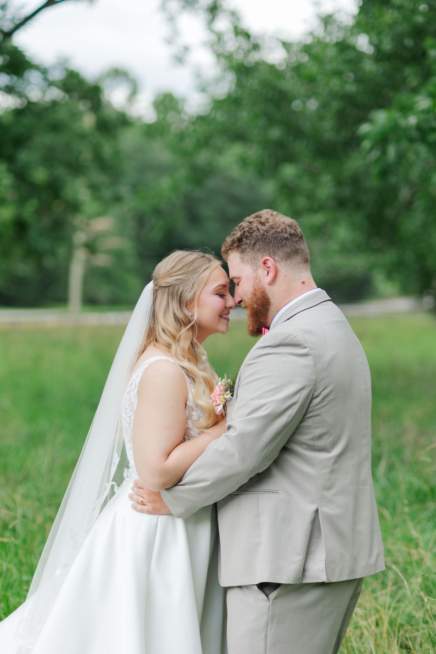 Bride and groom touch foreheads tenderly in a lush green outdoor setting, bride wearing a white gown and veil.