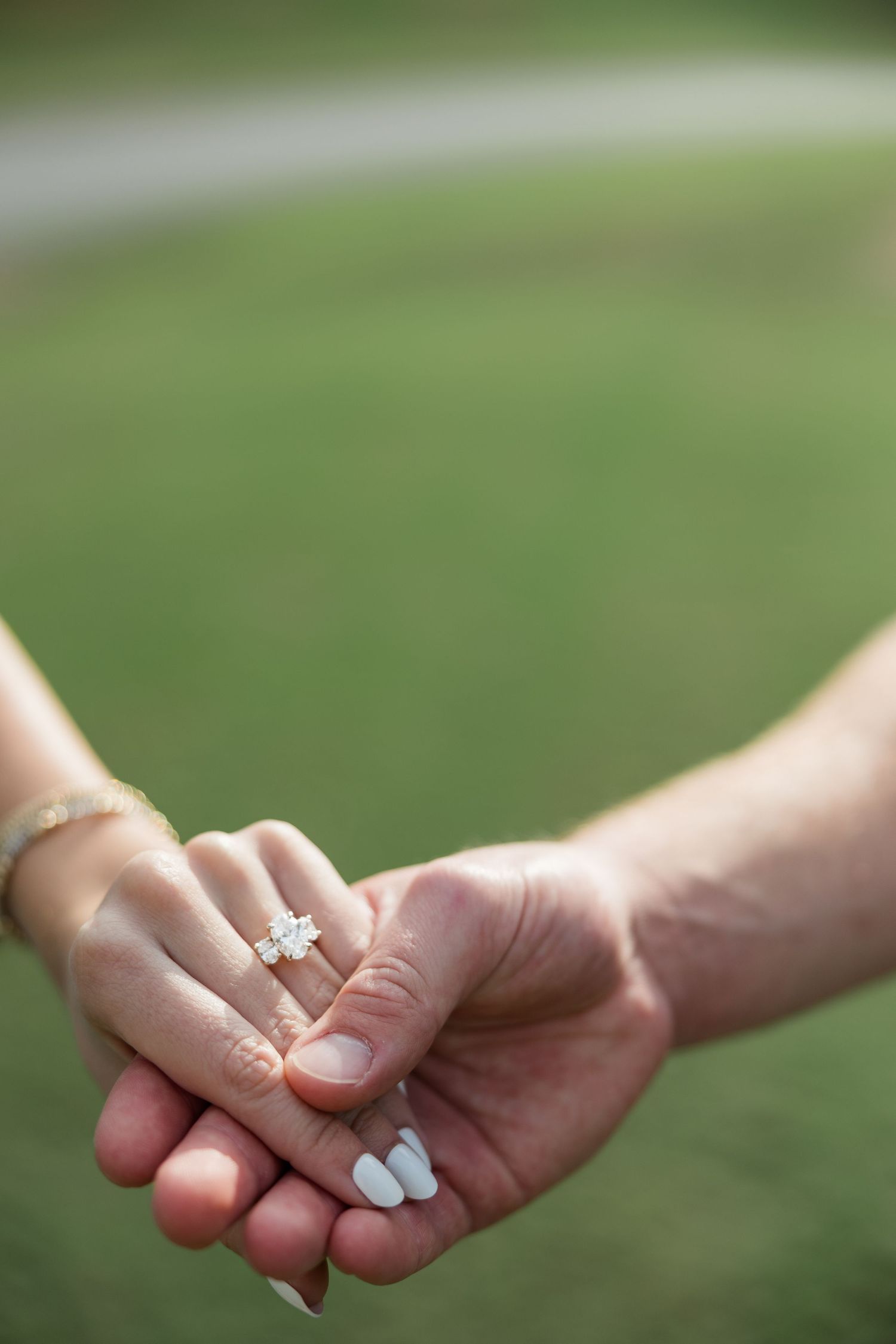 Close-up of a couple holding hands with a sparkling diamond engagement ring on a green grass background.
