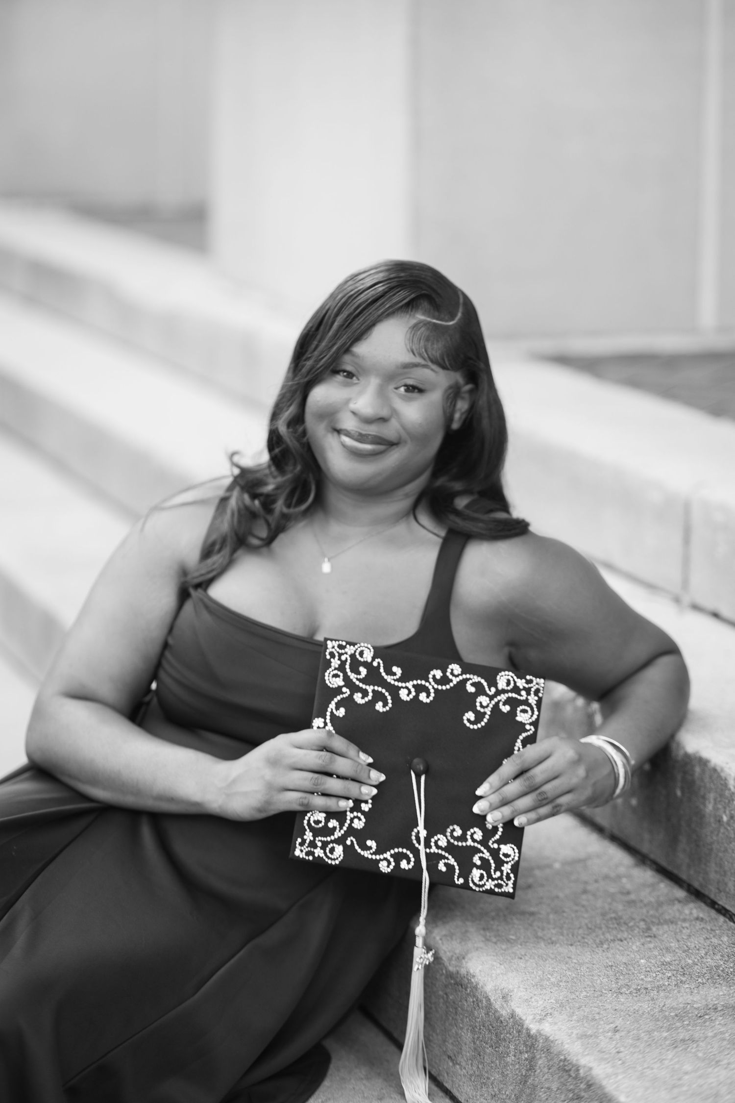 Black and white photo of a smiling young woman in a dress holding a decorated graduation cap on steps.