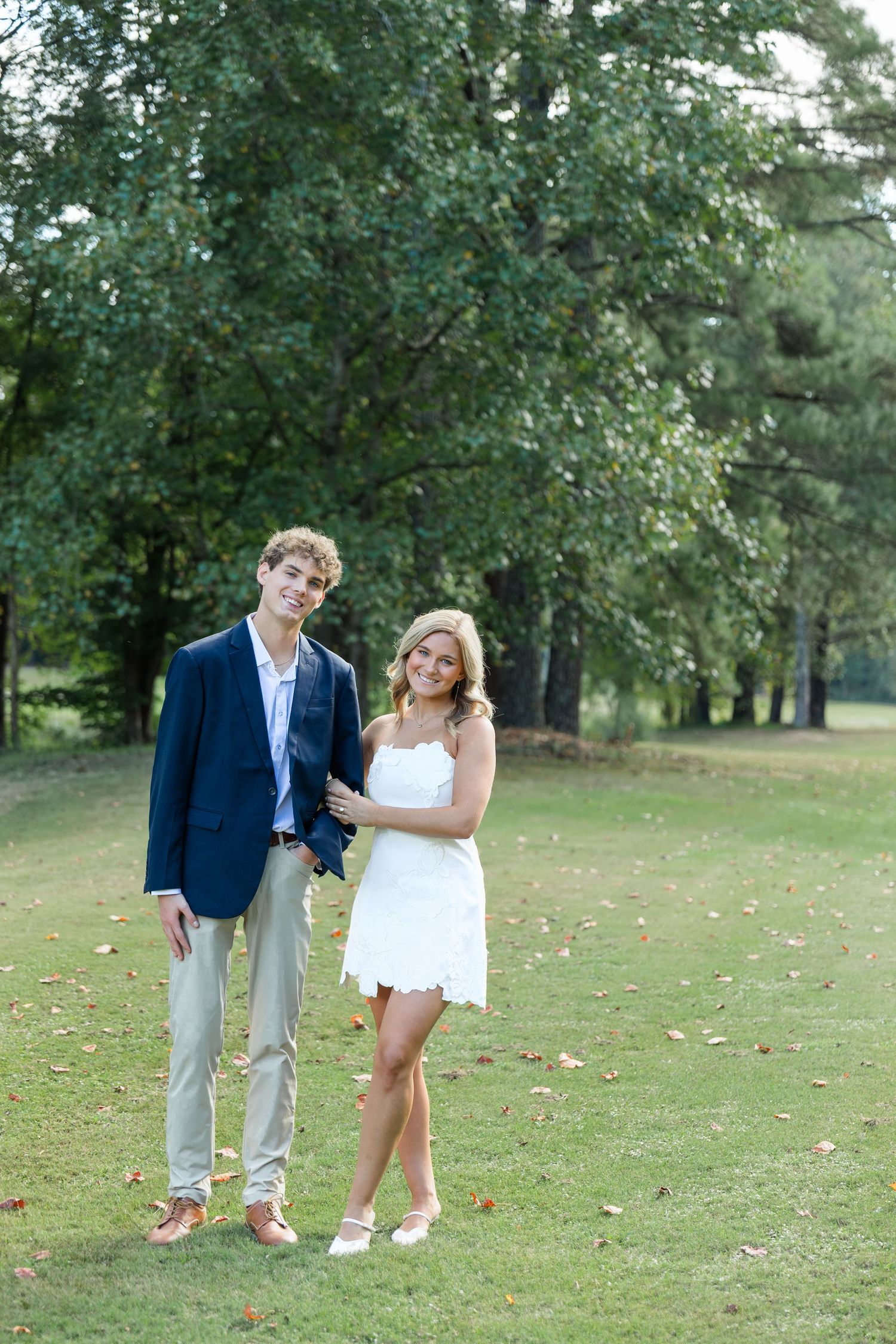 Young couple posing outdoors on golf course, man in navy blazer, woman in white mini dress.
