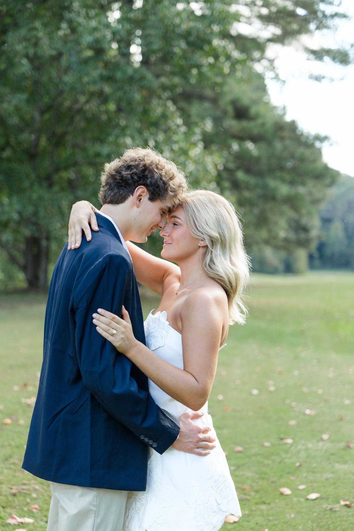 A couple in wedding attire embraces tenderly outdoors, the bride in white and groom in navy suit, surrounded by lush green trees.