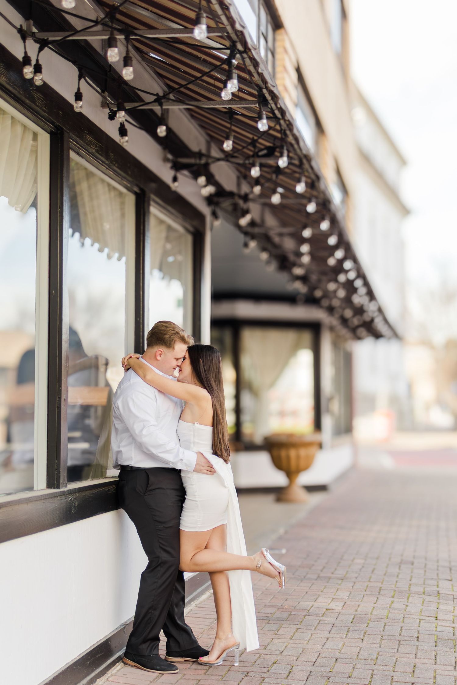 Couple embracing and kissing outside a restaurant with string lights, woman in white dress lifting her leg.