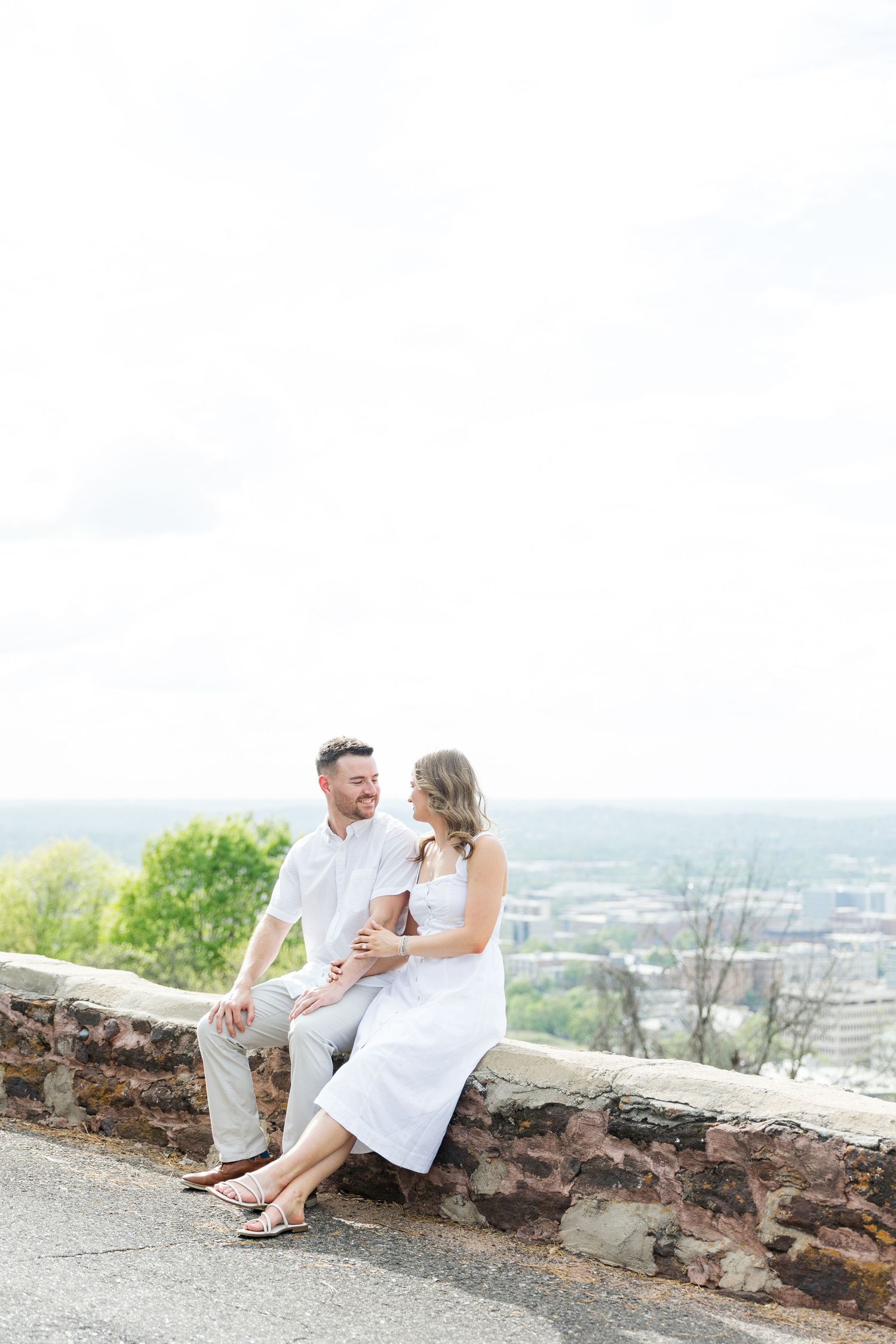 A couple in white sits on a stone wall overlook, smiling at each other with a city skyline in the background.