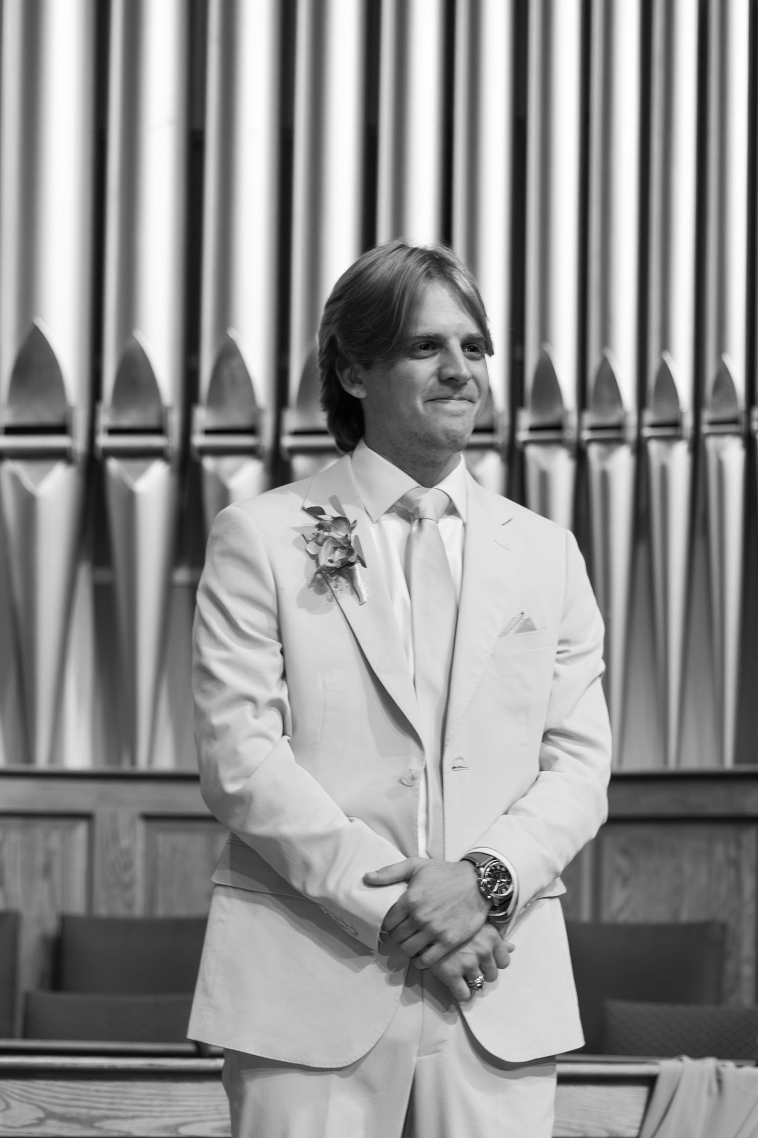 Black and white portrait of a smiling groom in a white suit standing before church pipe organ.