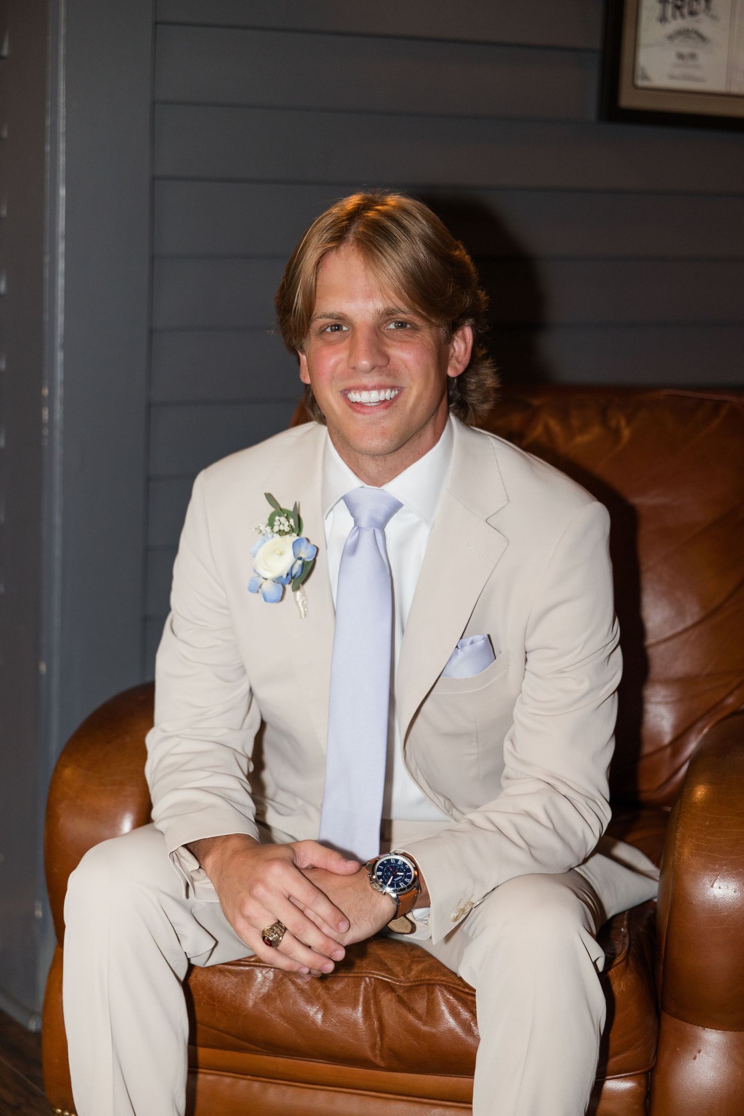 Smiling groom in tan suit with blue tie and boutonniere sitting in leather chair at wedding.