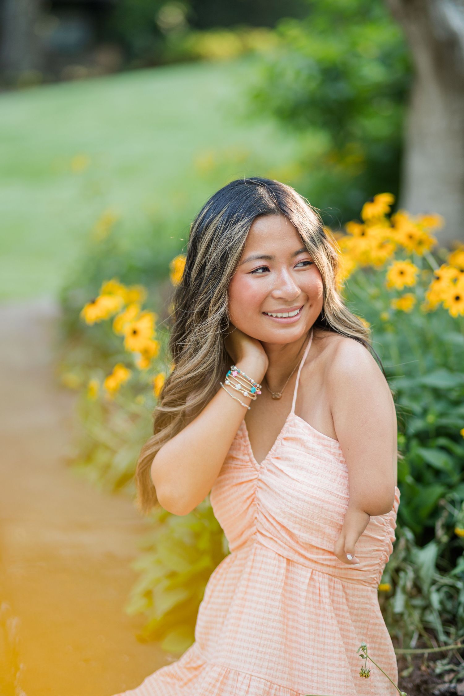 Cheerful young woman in a peach gingham dress posing beside bright yellow flowers in a garden.