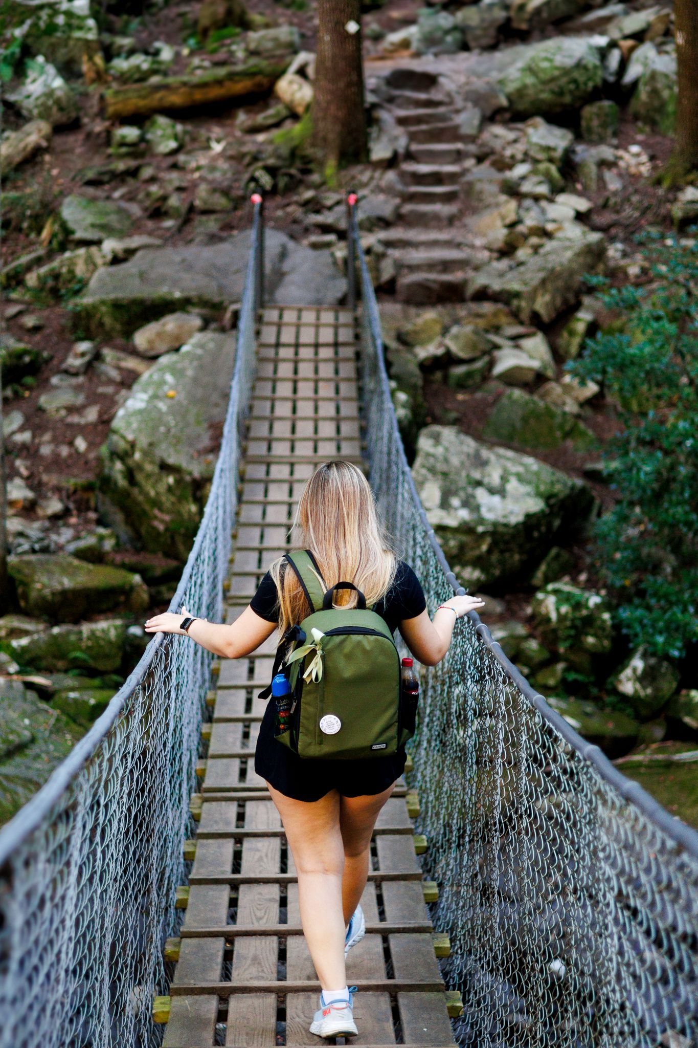 Blonde woman with green backpack crossing a wooden suspension bridge toward rocky stone steps.